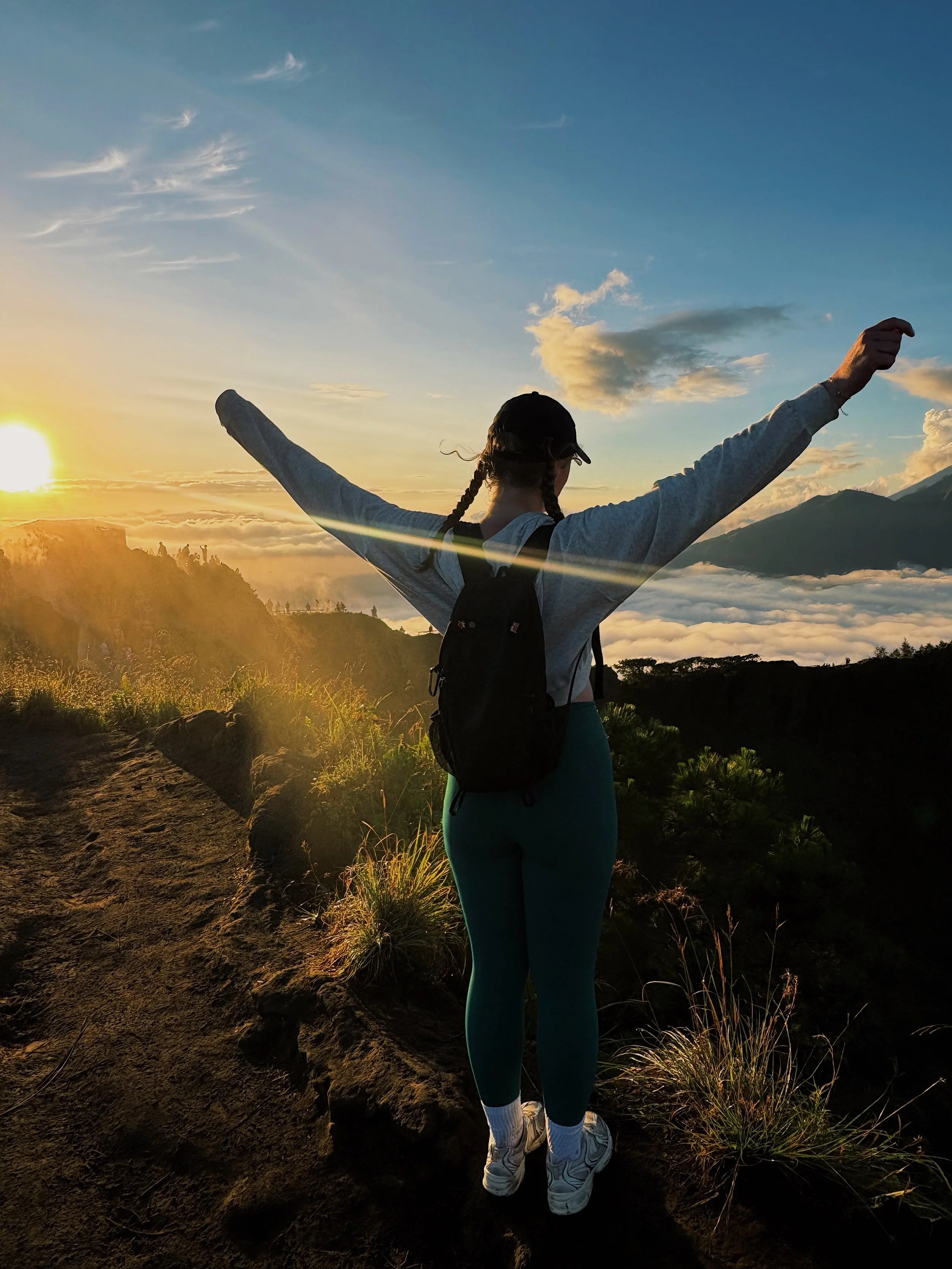 Person standing on a mountain trail with arms raised during sunset, overlooking clouds and distant mountains.