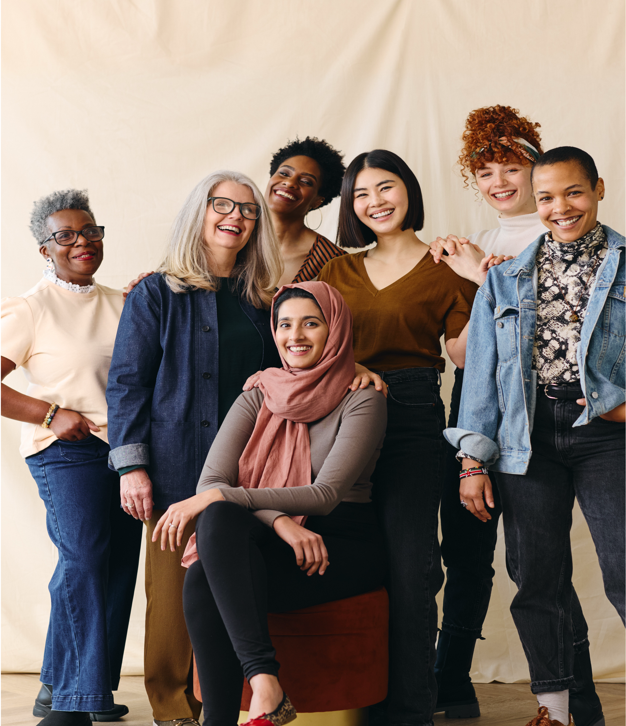 A diverse group of seven women standing and sitting in front of a plain background, smiling at the camera
