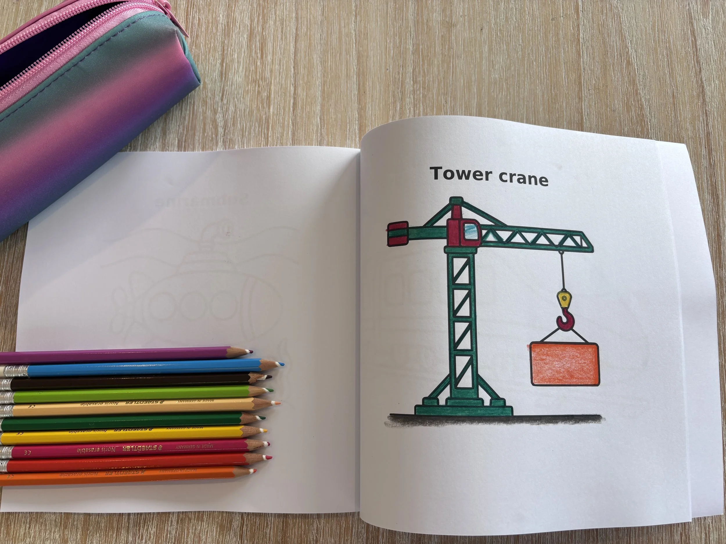 Open coloring book showing a tower crane lifting a red container, with colored pencils and a pink pencil case on a wooden table.