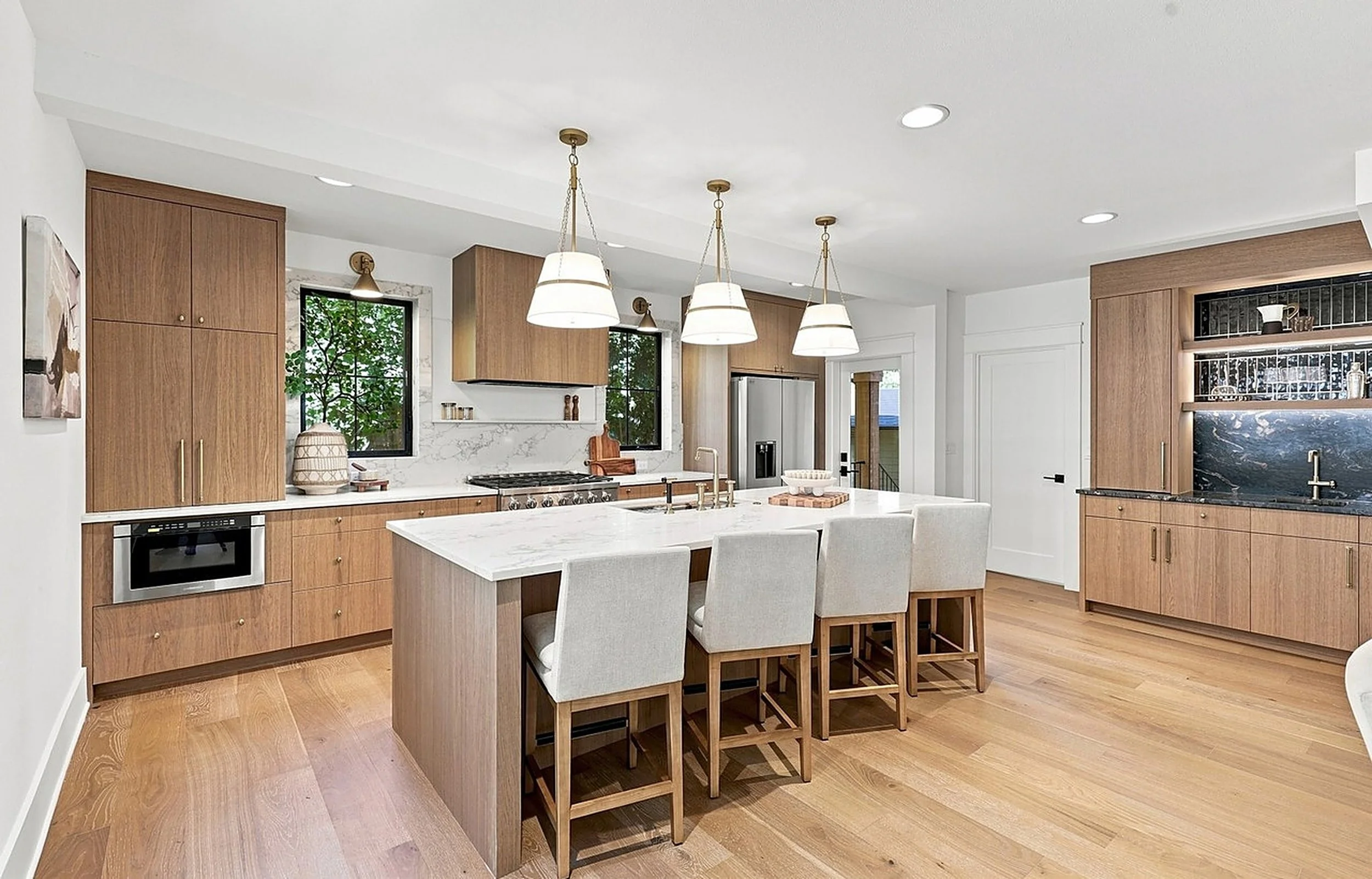 Staged kitchen with a long island, three pendant lights, cream counter stools and warm wood cabinetry