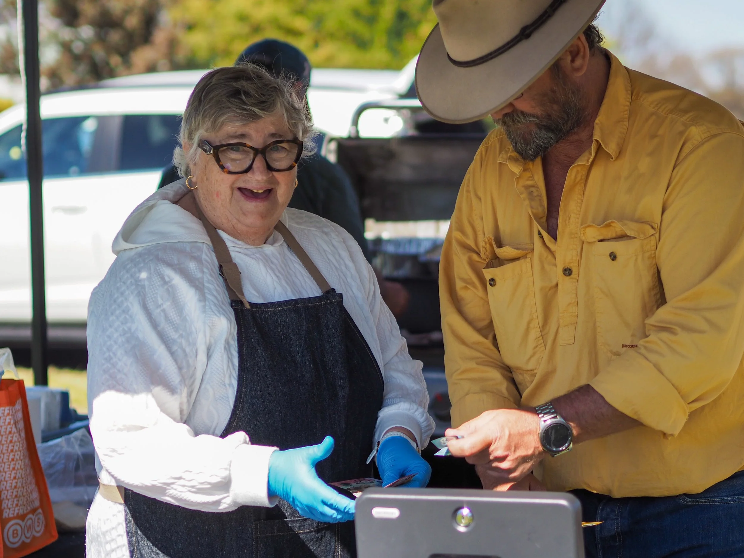 A woman wearing glasses, a white shirt, a dark apron, and blue gloves is receiving assistance from a man with a beard, glasses, a tan hat, and a yellow shirt during an outdoor event. They are working together, possibly preparing or serving food.