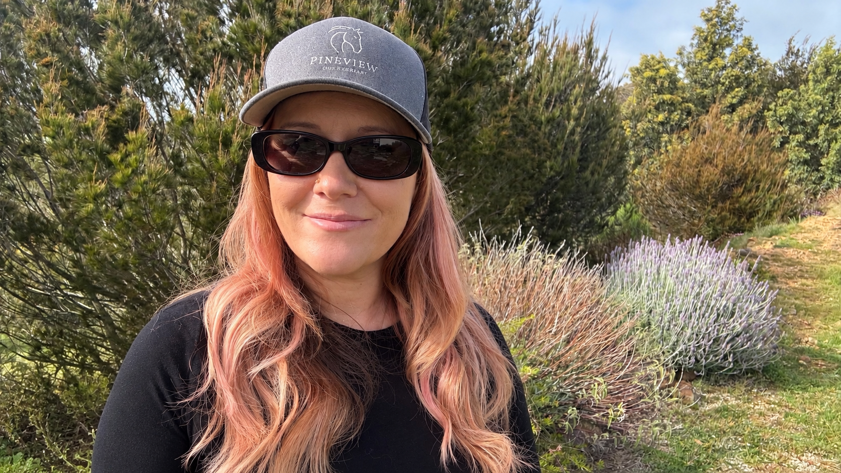 Woman with long pinkish hair wearing sunglasses and a gray cap with 'Pineview' logo, standing outdoors with green bushes and purple foliage in the background.
