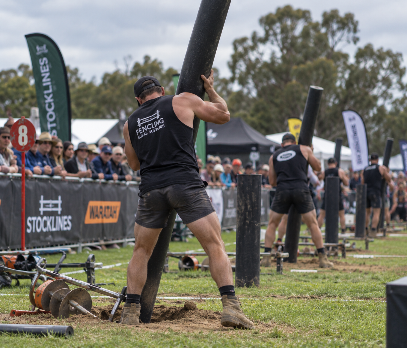 Men participating in a log lifting competition outdoors, with spectators watching behind safety barriers.