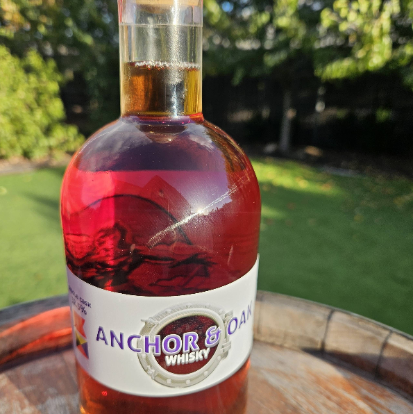 A bottle of Anchor & Oaken whisky on a wooden surface outdoors with a grassy yard and trees in the background.