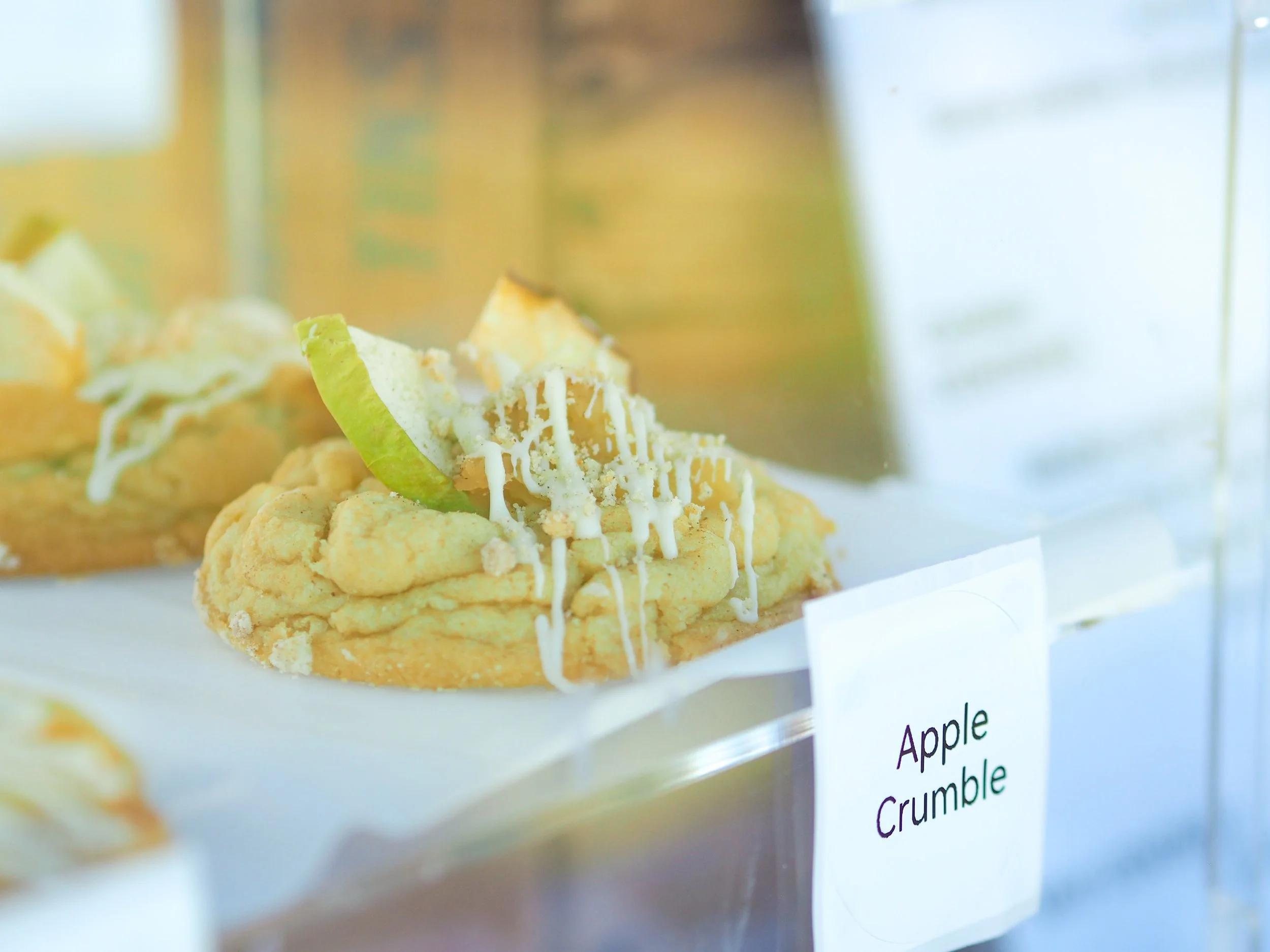 Close-up of apple crumble dessert with a green apple wedge and white drizzle topping, displayed behind a glass case.