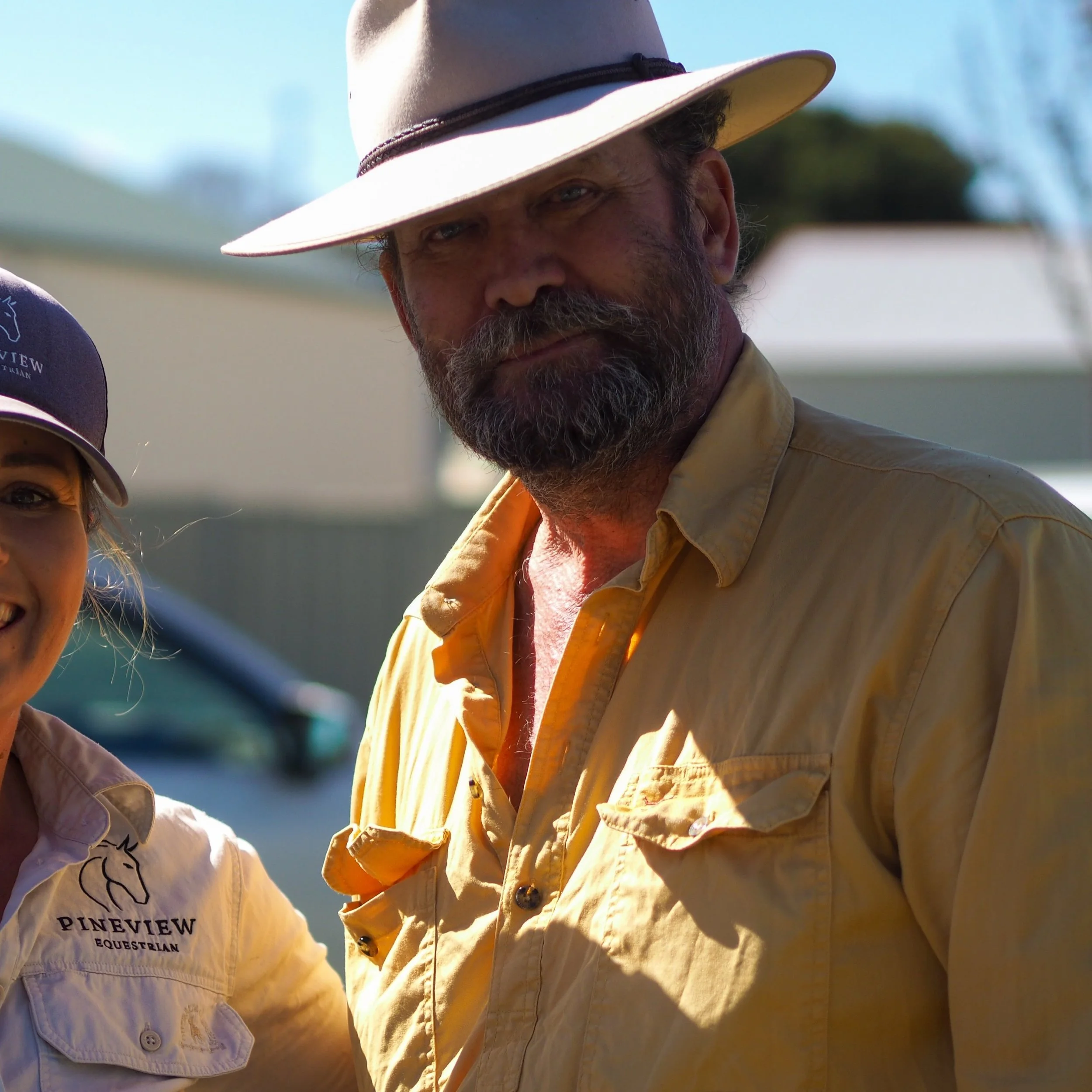Close-up of a man wearing a wide-brimmed hat with a black band, focusing on his face with a beard, outdoors with a blue sky and trees in the background, wearing a yellow shirt.