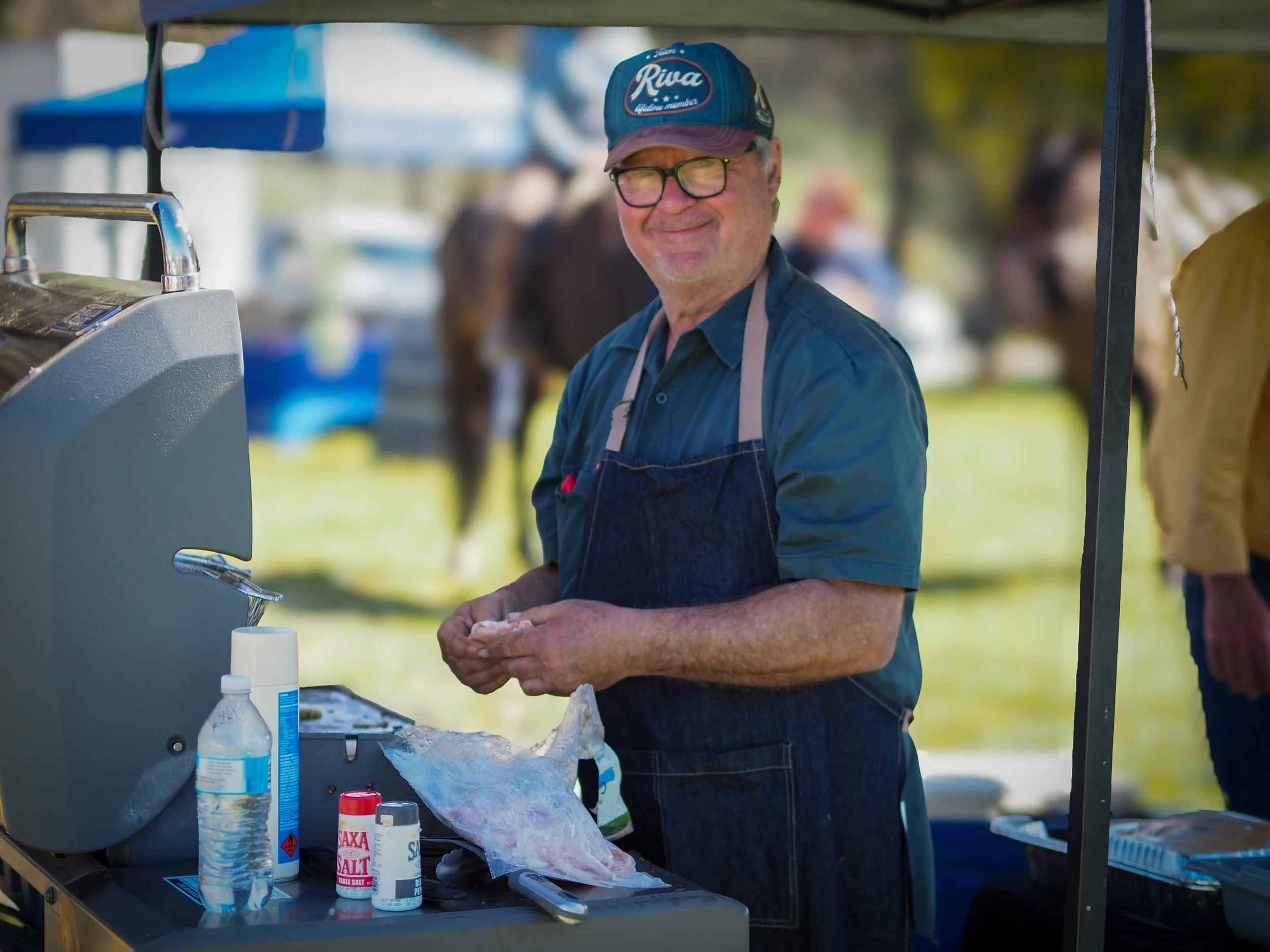 A smiling man with glasses and a blue cap stands at an outdoor food stand, preparing food with various ingredients and condiments on the counter.