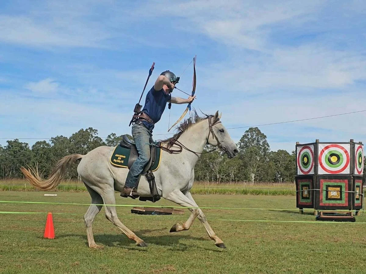 A man riding a white horse aiming a bow and arrow at an archery target outdoors on a grassy field.