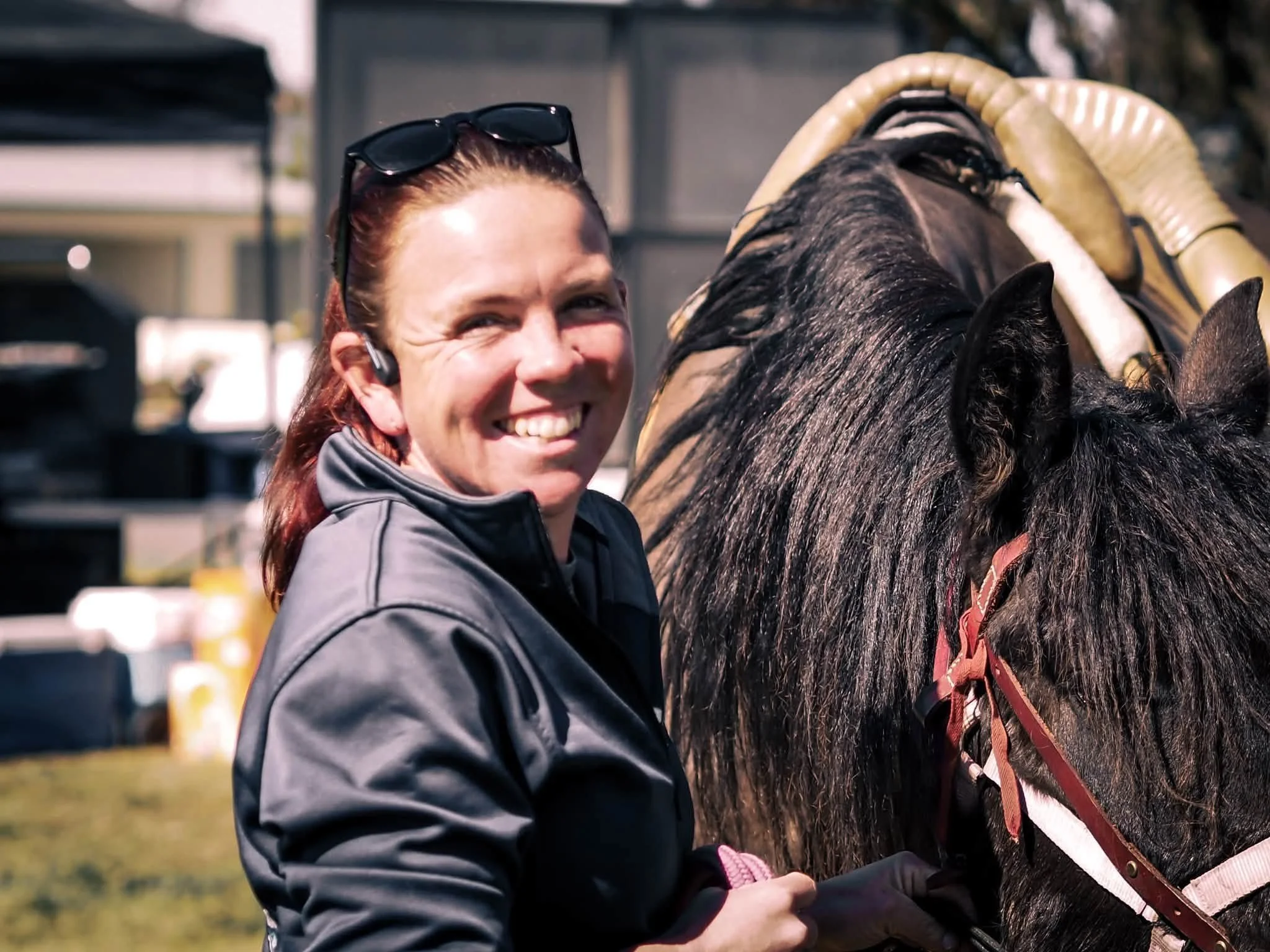 A woman smiling and wearing sunglasses on her head, standing next to a dark horse with a bridle, outdoors in a sunny setting.