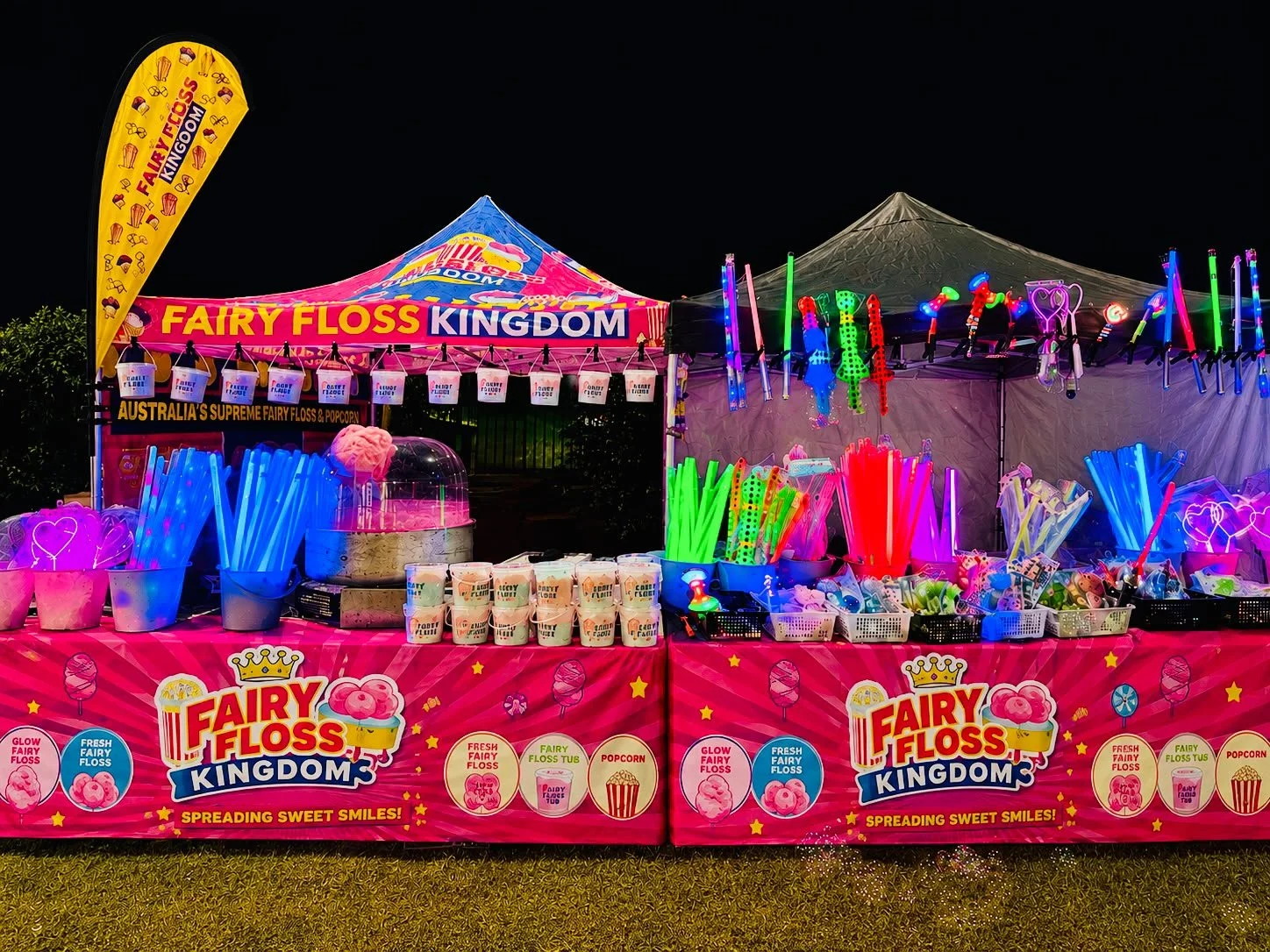 A brightly lit fairground stall at night selling fairy floss, popcorn, and fairy floss tubs. The stall has pink cloth with branding and colorful lights, with various glow sticks and toys hanging and displayed on the table and canopy.
