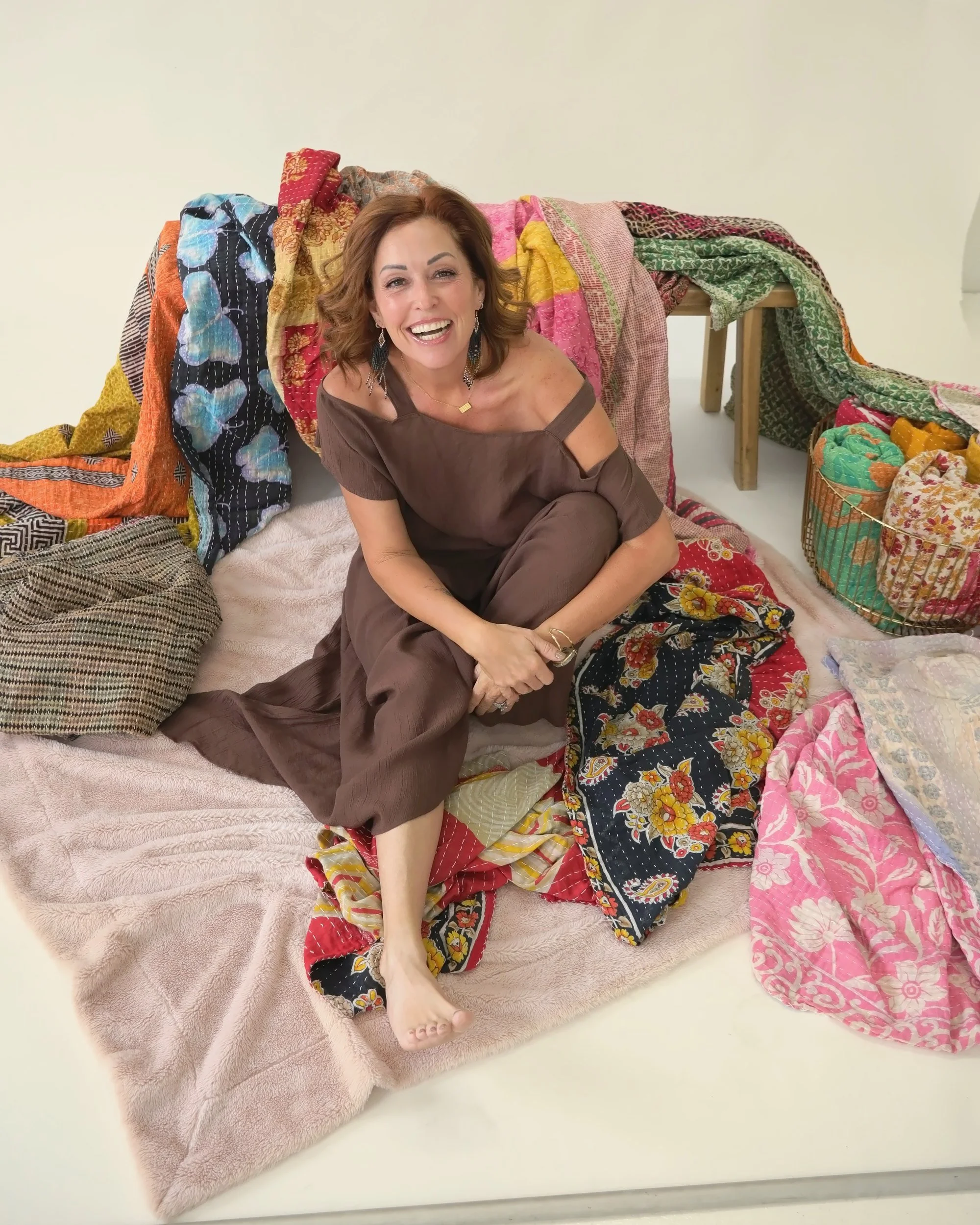 Wende Valentine smiling, sitting among a pile of colorful kantha blankets