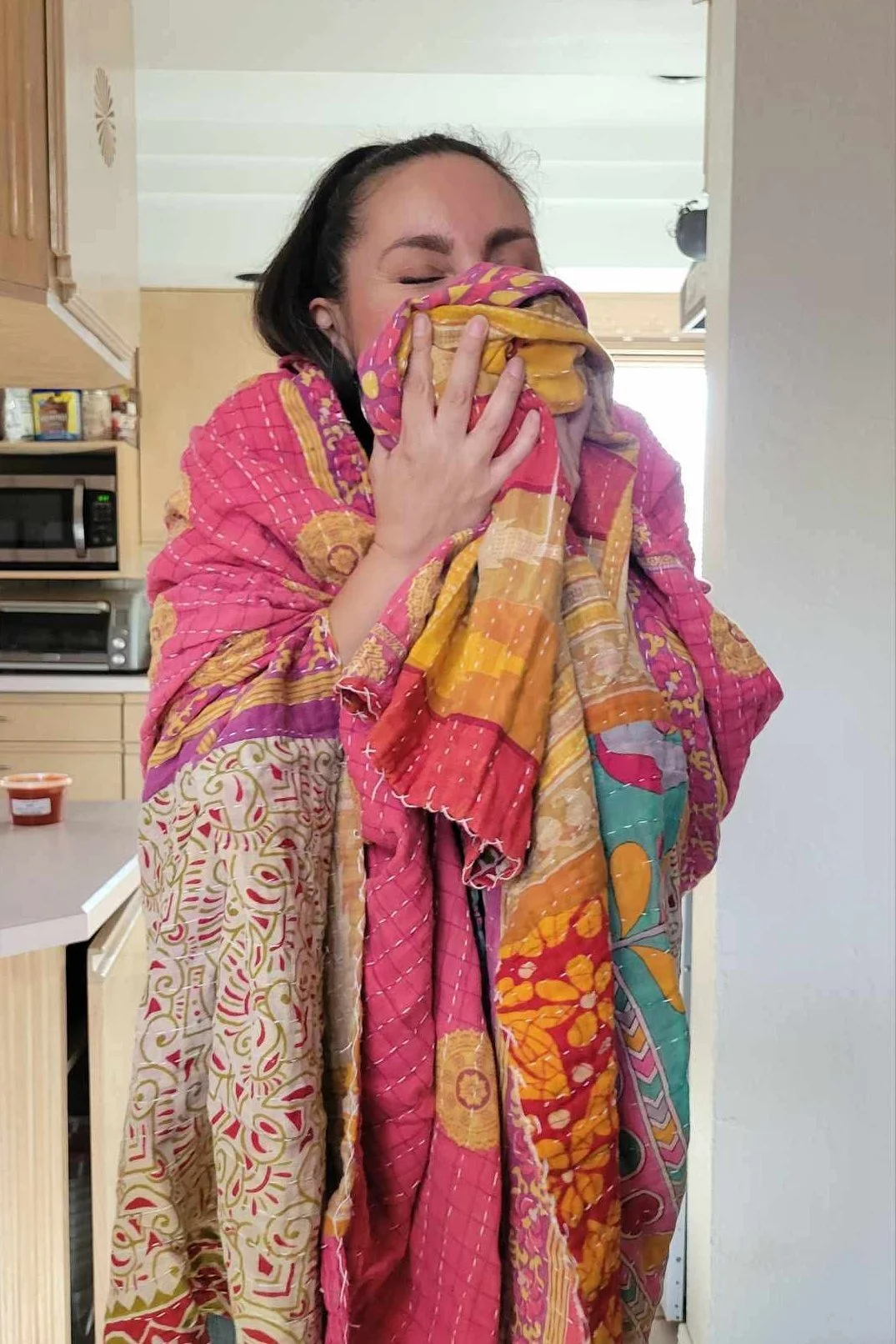 A woman wrapped in a pink kantha blanket, holding it to her face with her eyes closed