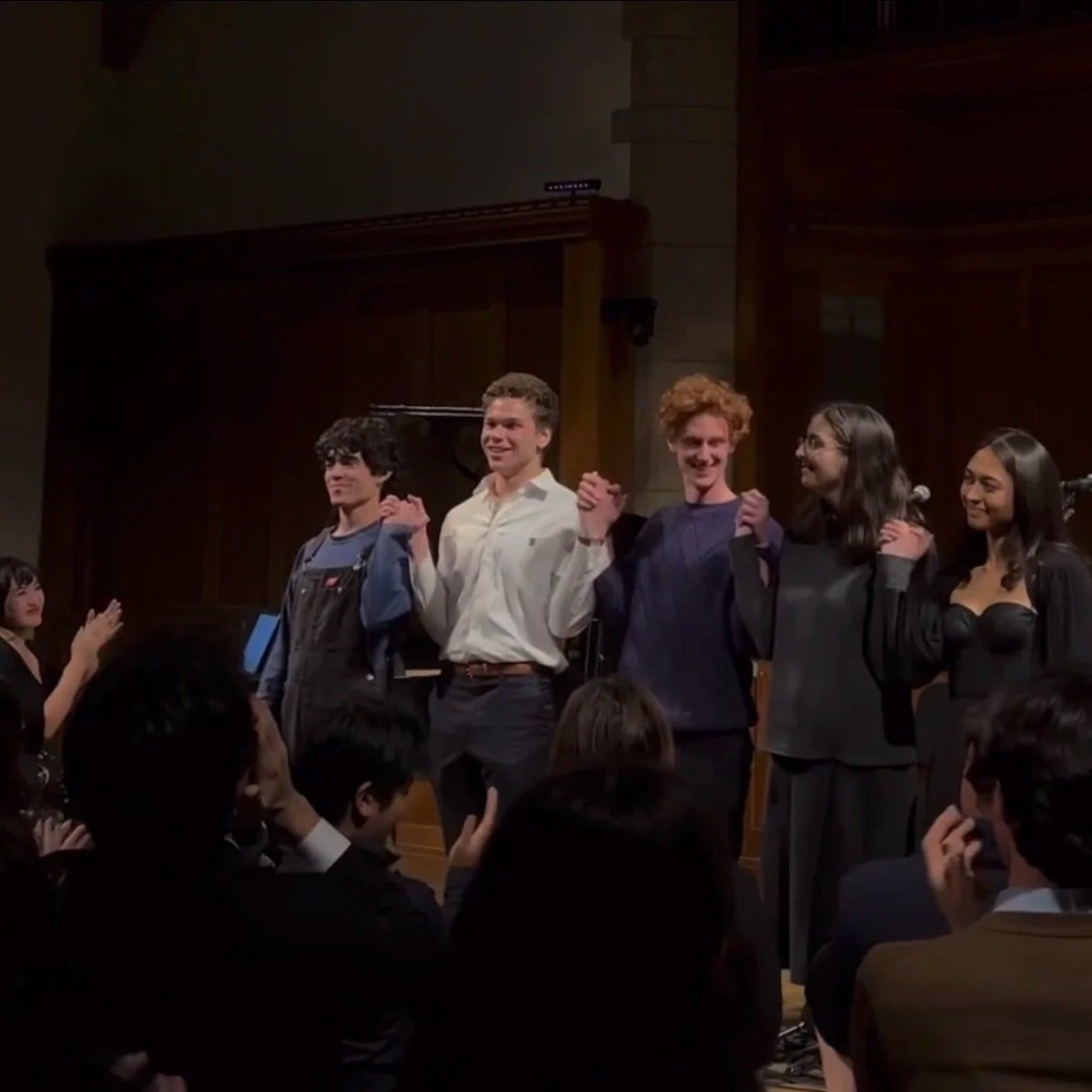 Group of six young adults on stage holding hands and smiling, with audience members in front applauding and taking photos.