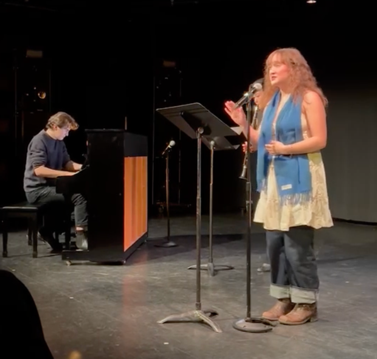 A woman singing into a microphone on stage, with a pianist playing a black piano nearby, in a dimly lit performance space.