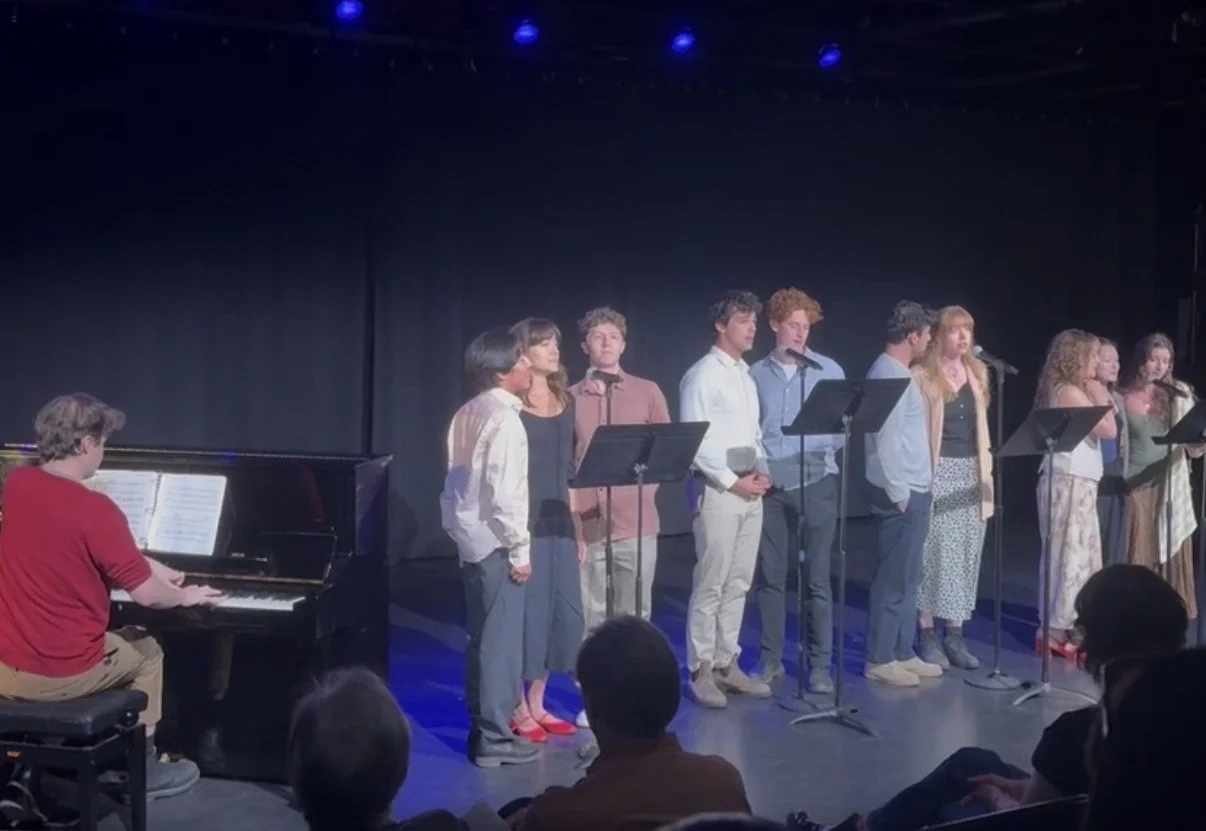 A group of nine young singers performing on stage with a pianist accompanying them, under stage lighting, with an audience watching.