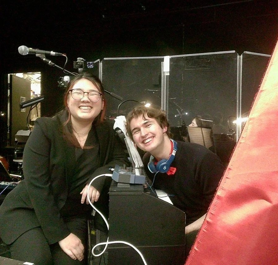 Two people smiling in a recording studio, one is sitting at a piano with headphones and the other is standing beside her. There are microphones and electronic equipment around them.