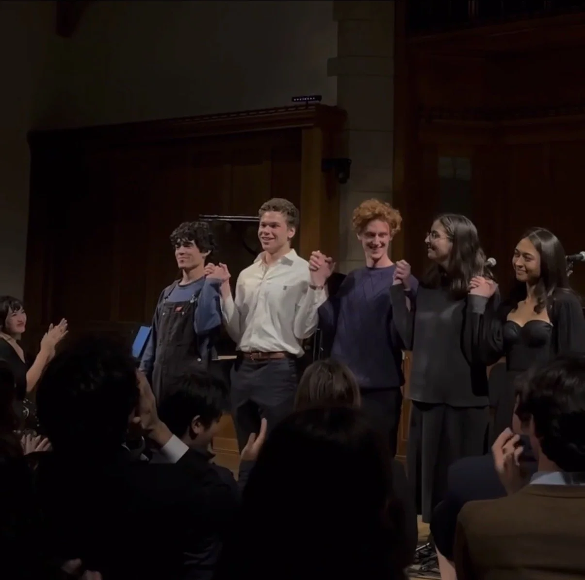 Group of five people holding hands and taking a bow on stage during a performance or ceremony, with an audience in front.
