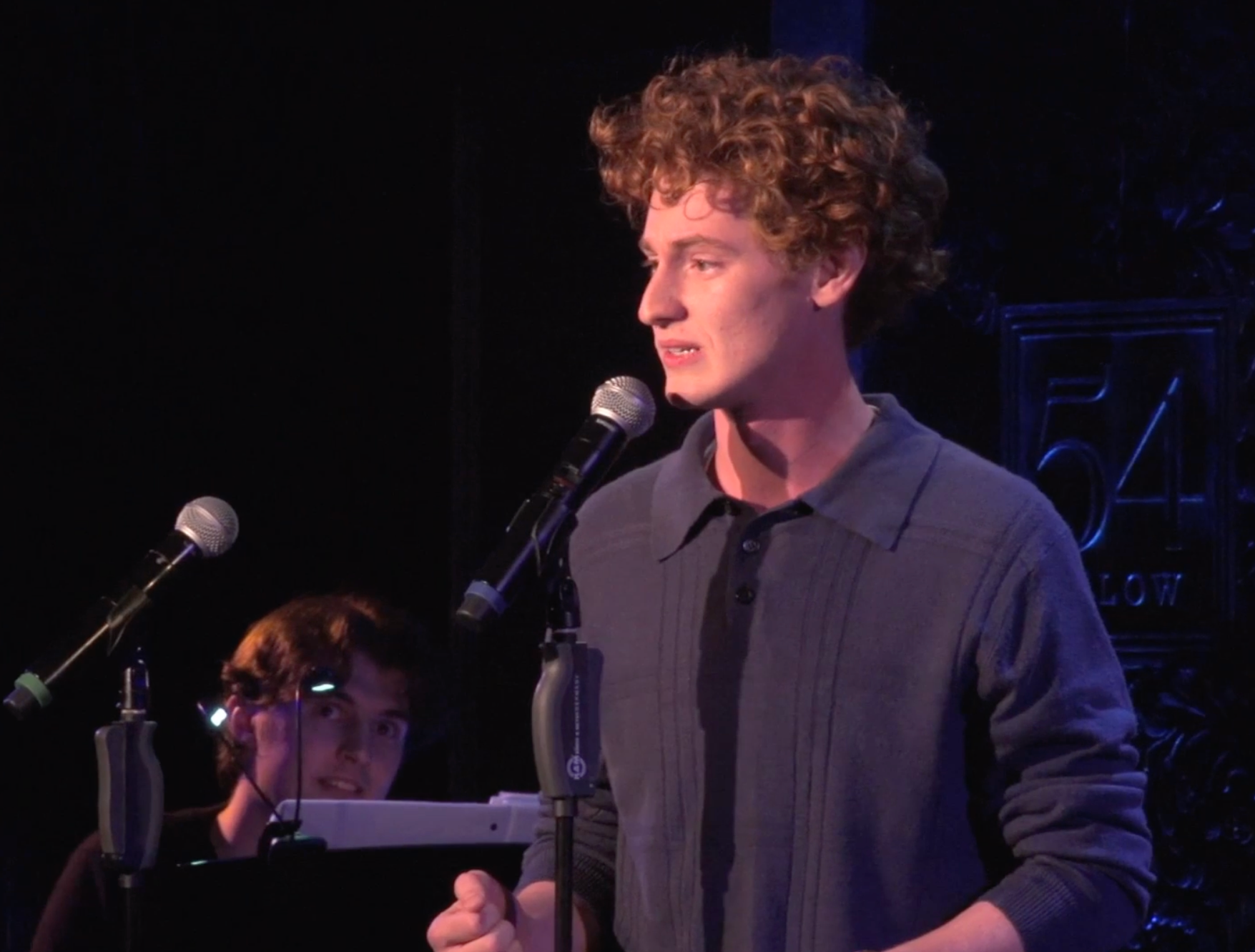 A young man with curly red hair stands at a microphone on a stage, speaking or singing, with a dark background and a sign that reads 'FOL...' in the background. Another person with glasses and headphones is visible in the background, seated at a keyboard.