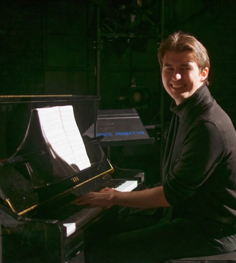 A young man smiling while playing a grand piano, with sheet music on the music stand, in a dark music studio or stage setting.