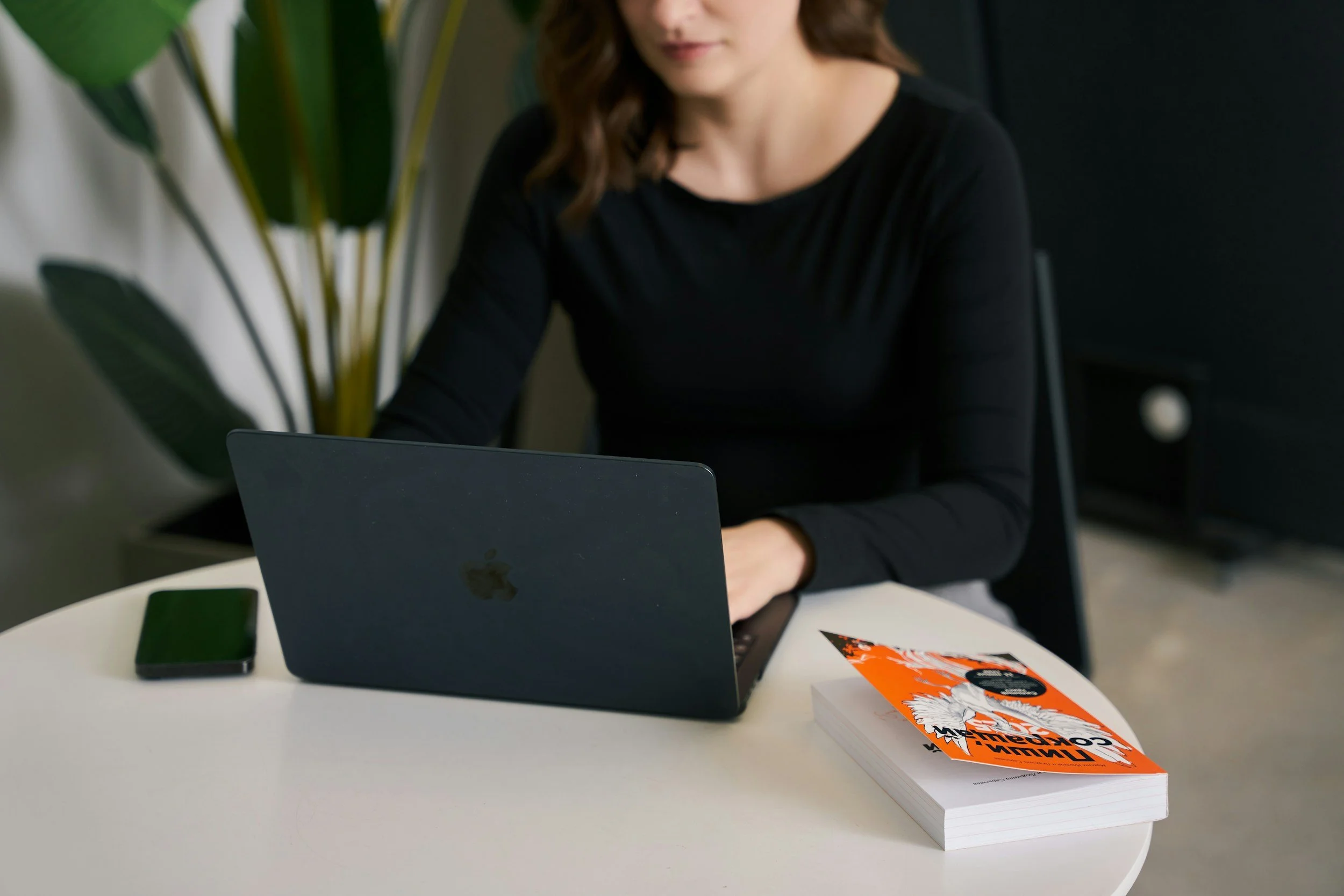 A woman sitting at a white table, working on a laptop. On the table, there is a black smartphone, a orange and black comic book titled 'FALLEN COMIX,' and a stack of papers or notebooks.