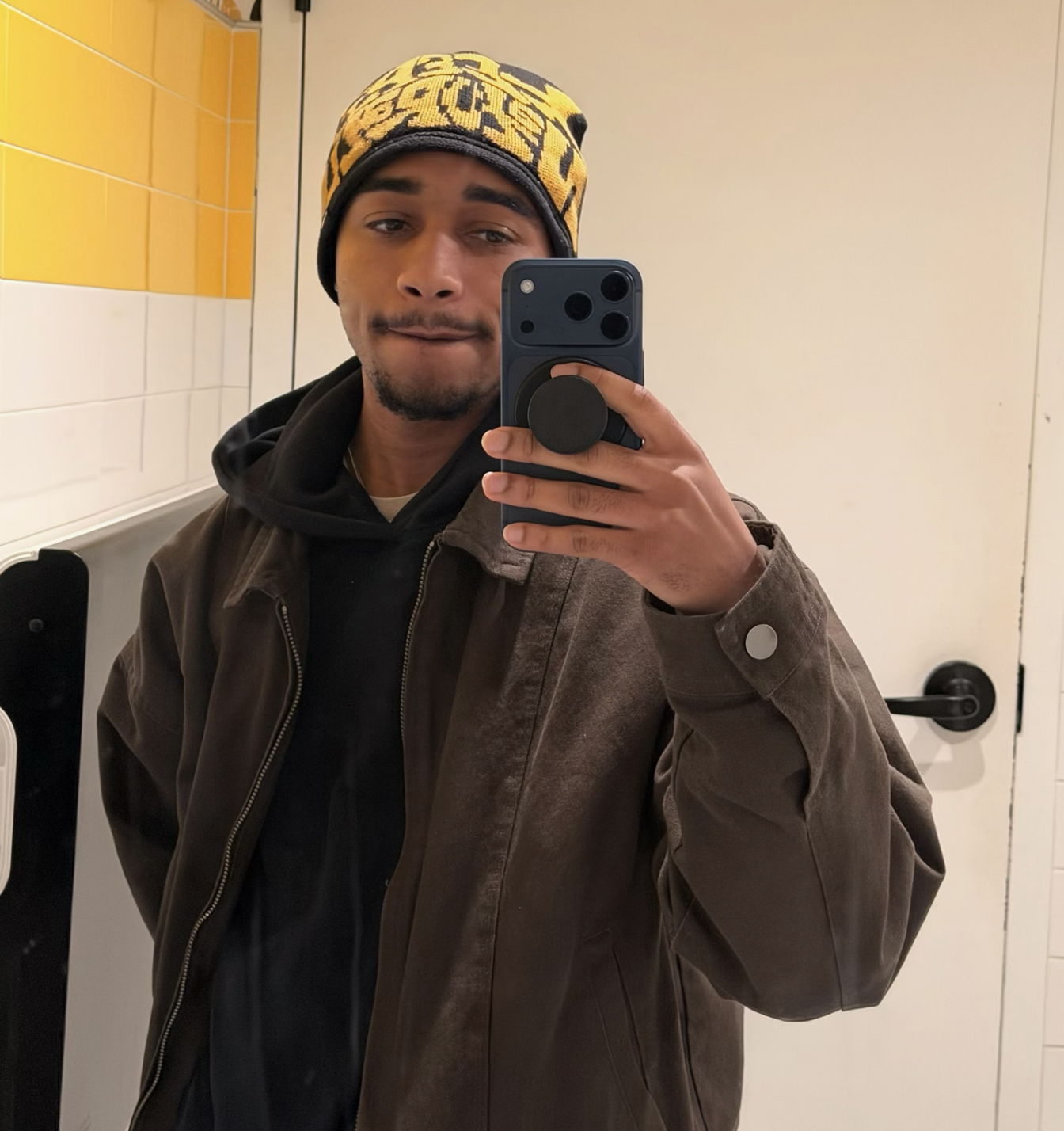 Rosiah wearing a patterned beanie and brown jacket taking a mirror selfie with a smartphone in a tiled bathroom