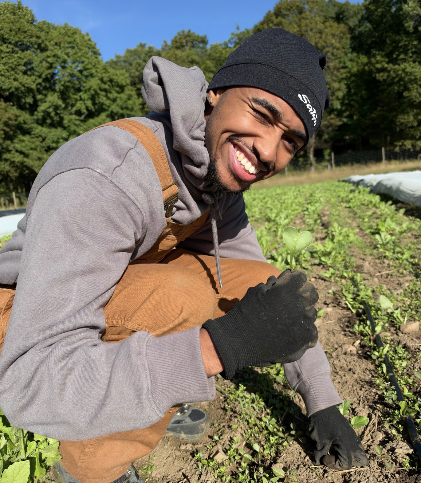 Smiling farmer in a beanie, hoodie, overalls, and gloves tending a young seedling in a field