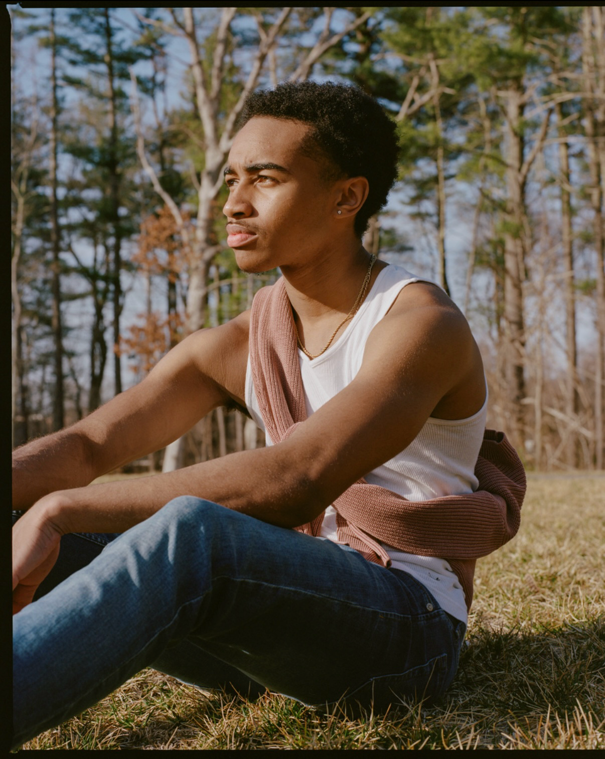 Rosiah sitting outdoors in a sunlit forest wearing a white tank top, blue jeans, and a brown sweater