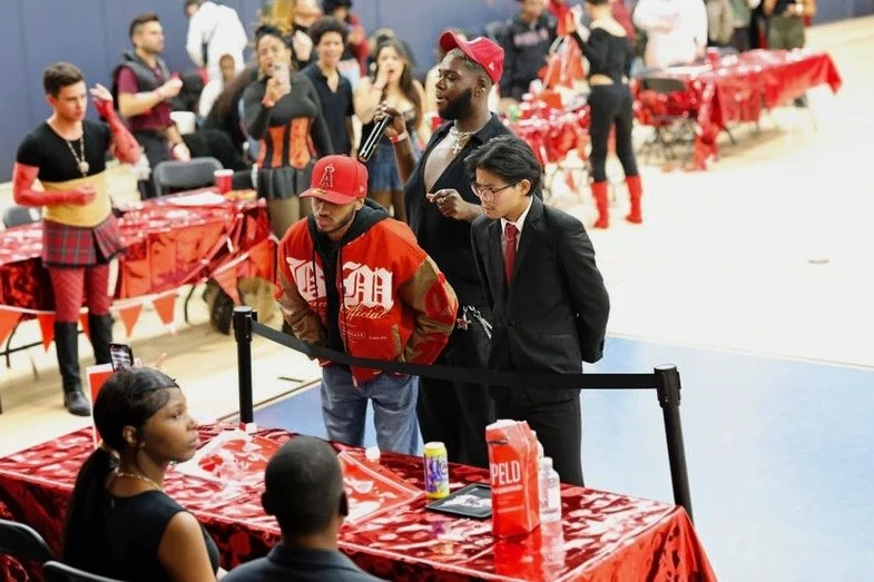 Rosiah in a red jersey and cap signing autographs at a crowded event table while supporters and judges look on