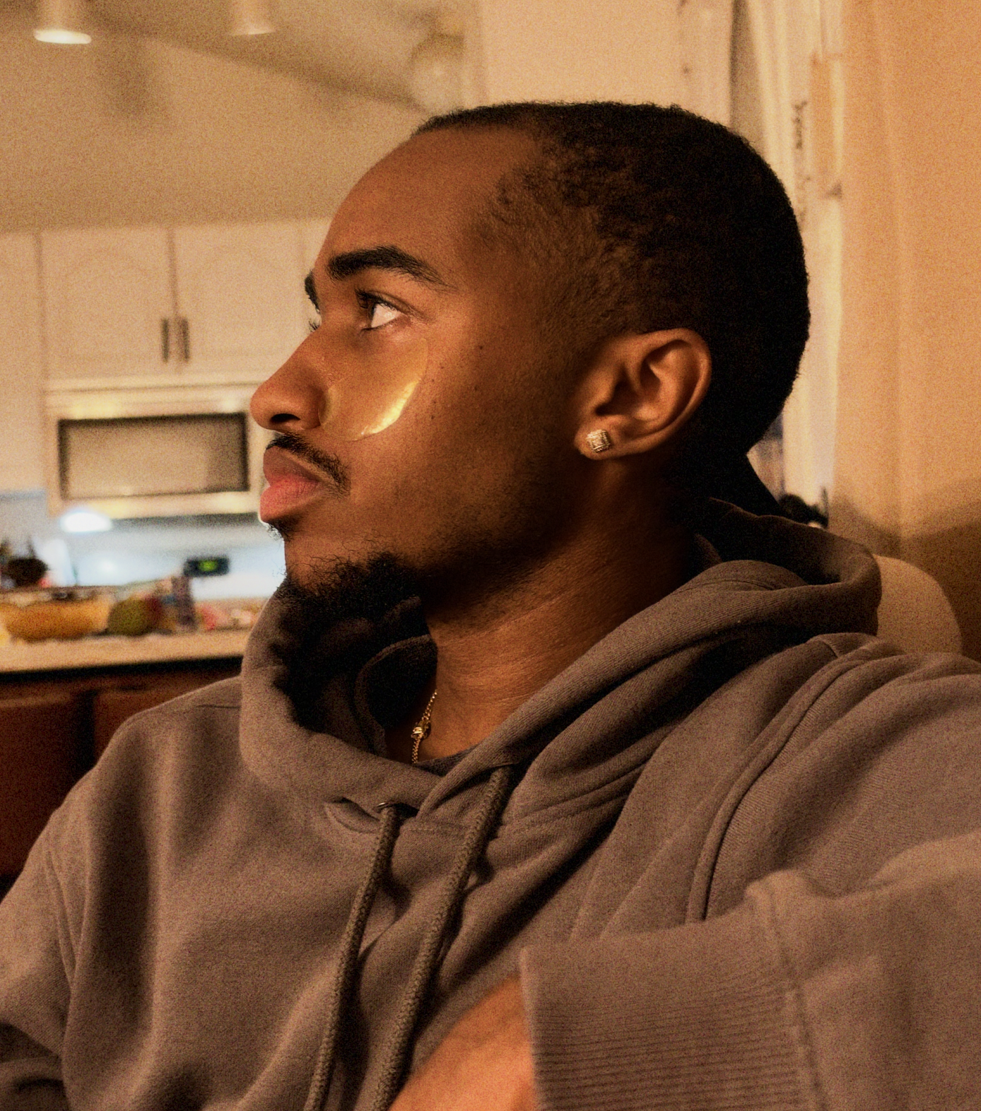 Side-profile portrait of Rosiah wearing a gray hoodie and square stud earring in a warmly lit kitchen