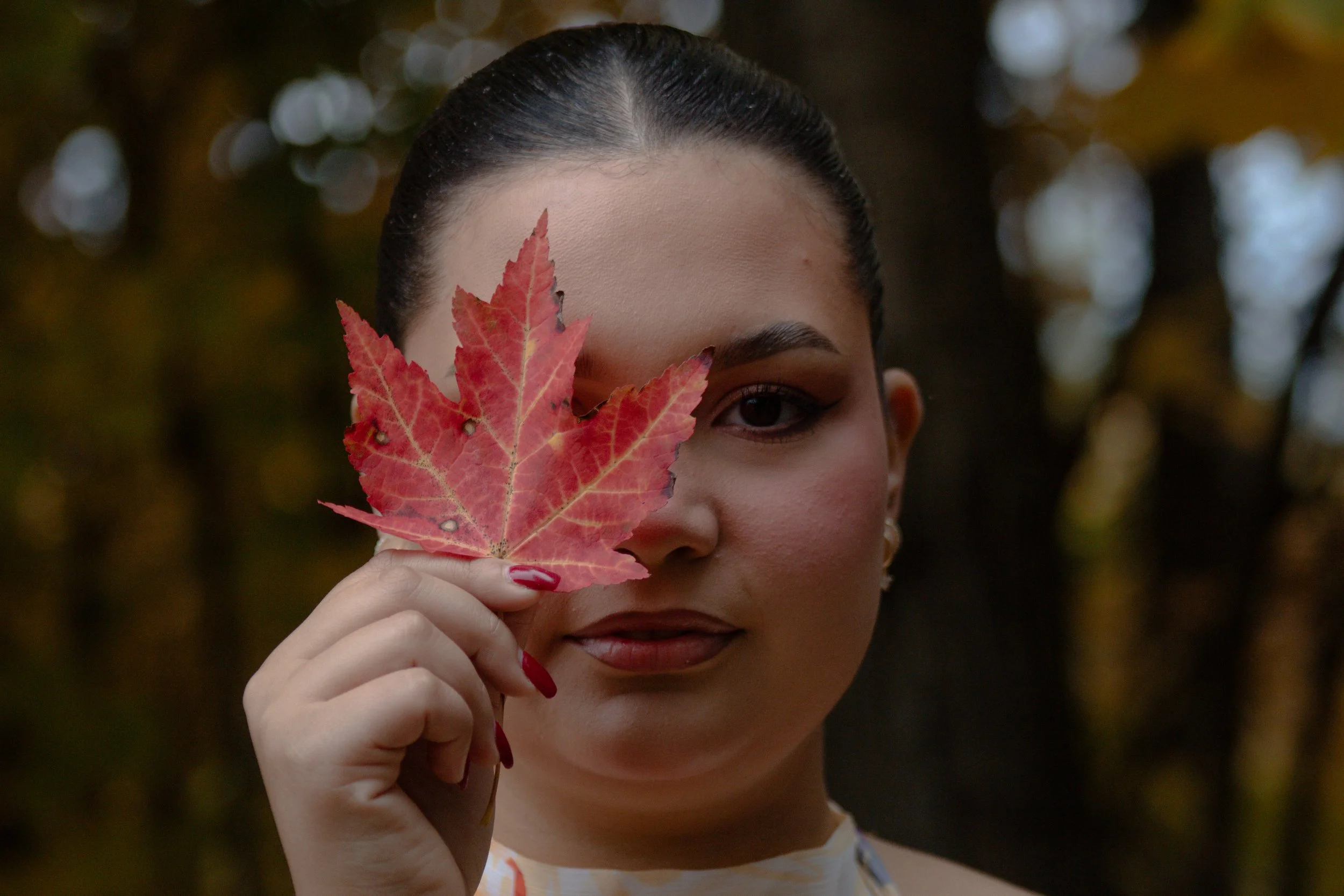 A woman with dark hair holds a red maple leaf in front of her face, covering her nose and mouth, with only one eye visible.