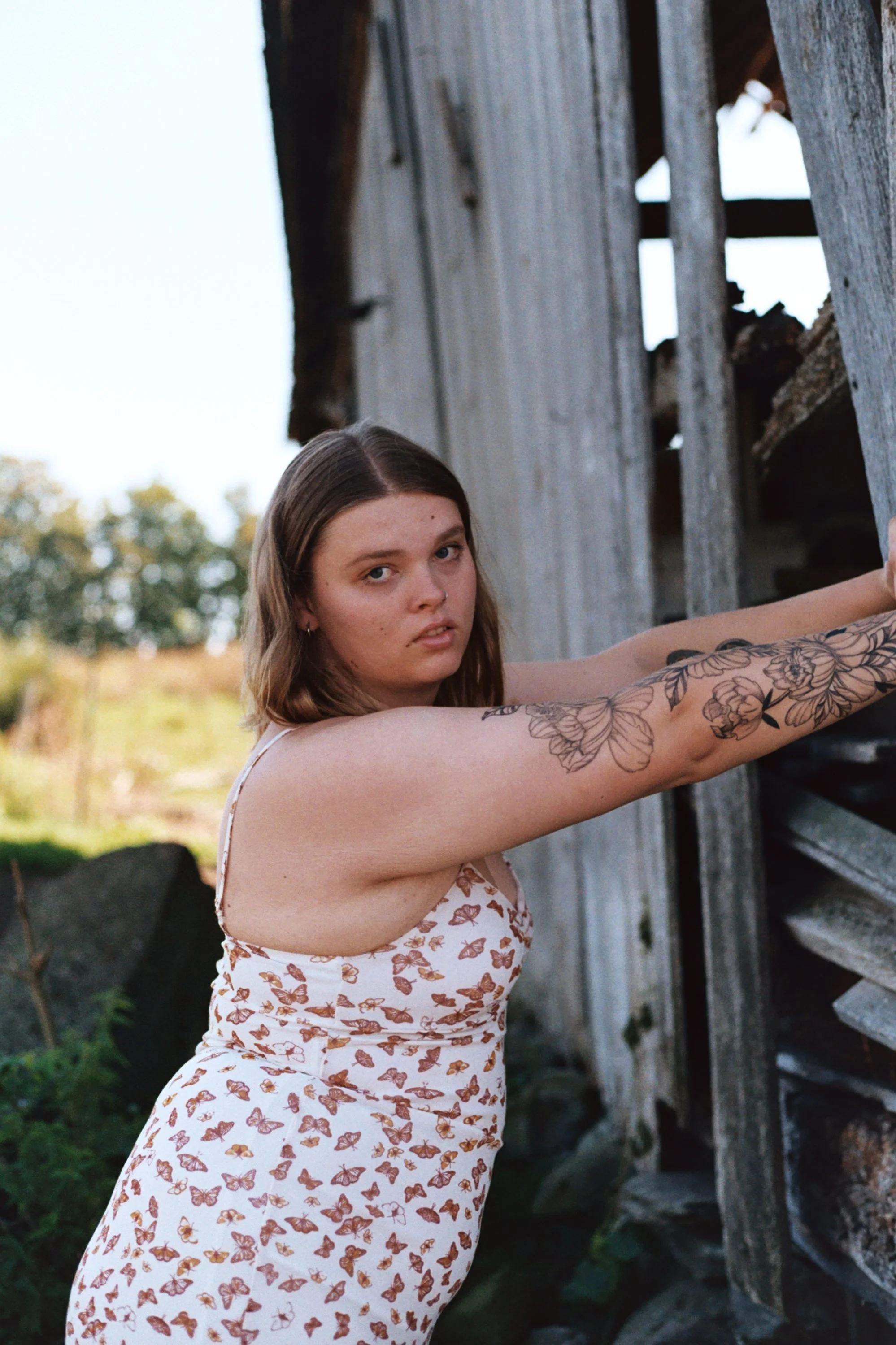 A young woman with medium-length brown hair and tattoos on her arm stands outdoors near a weathered wooden structure, wearing a white dress with a butterfly pattern, and looking at the camera.