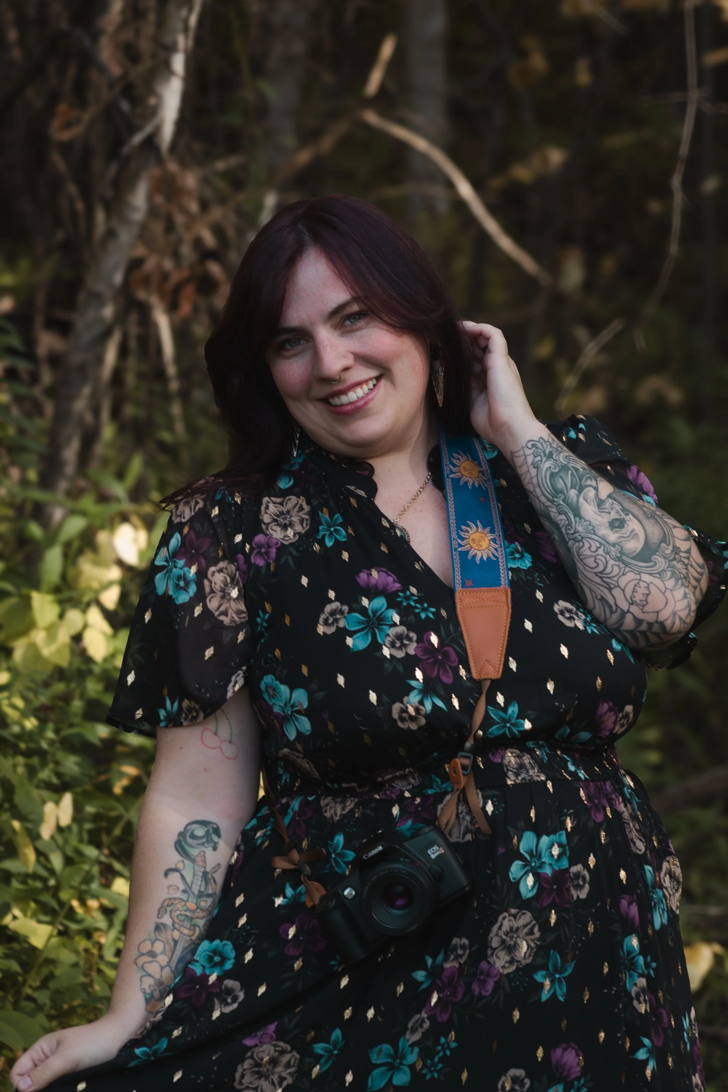 A woman with dark hair and tattoos smiling outdoors, wearing a floral dress and a camera around her neck, with trees and greenery in the background.