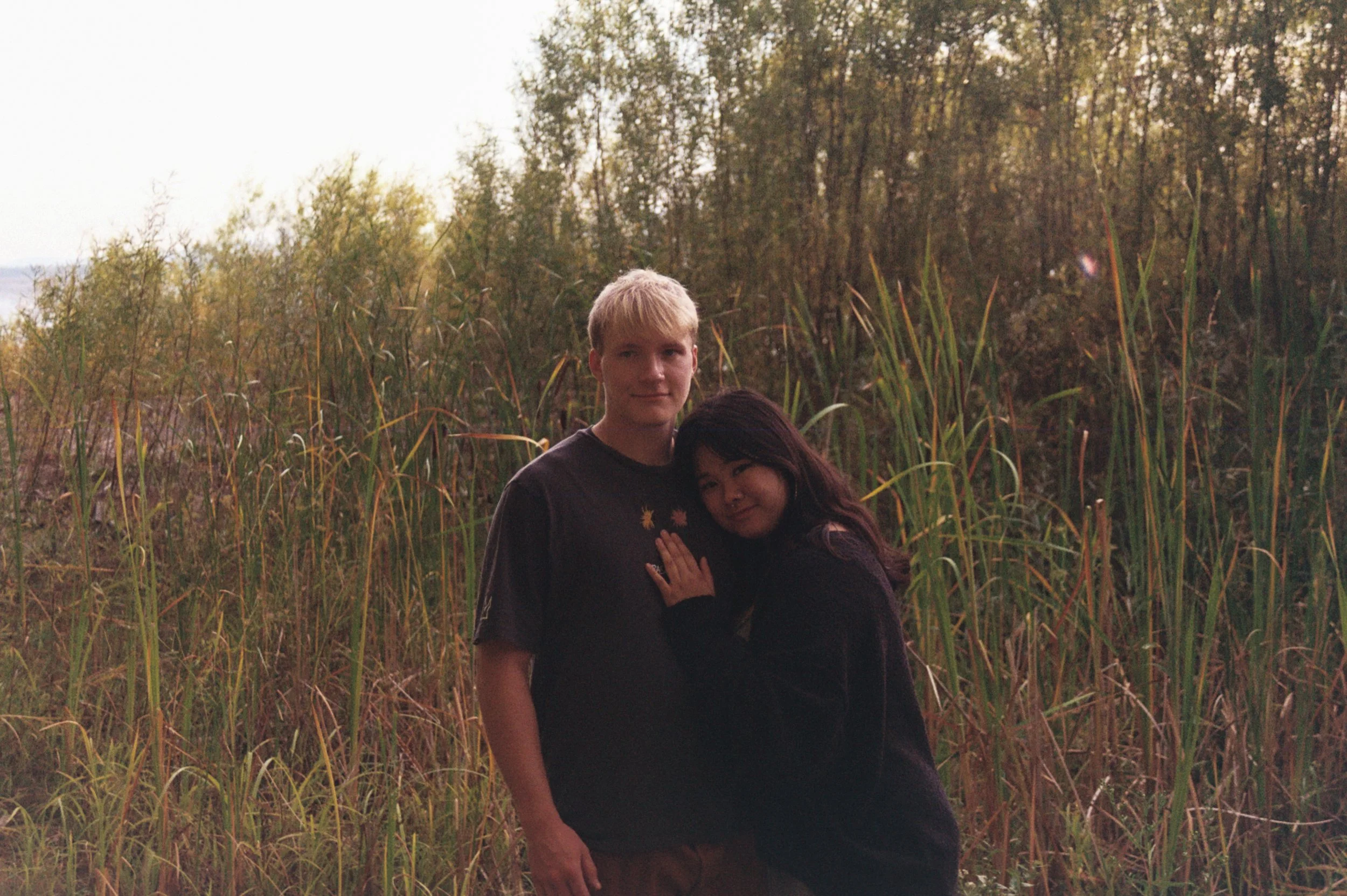 Two young people standing close together in front of tall grass and trees during sunset.
