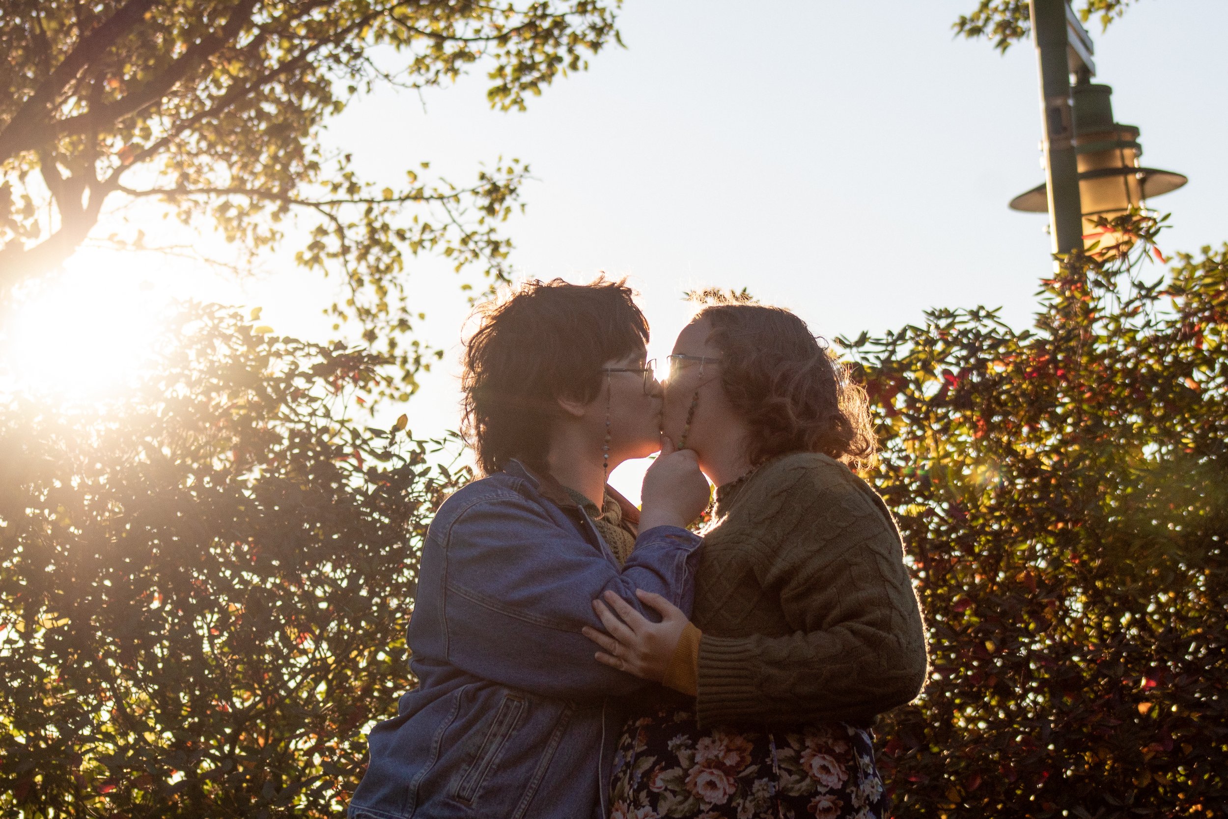 A couple kiss under the sunlight, surrounded by trees and leaves, with a lamp post in the background.
