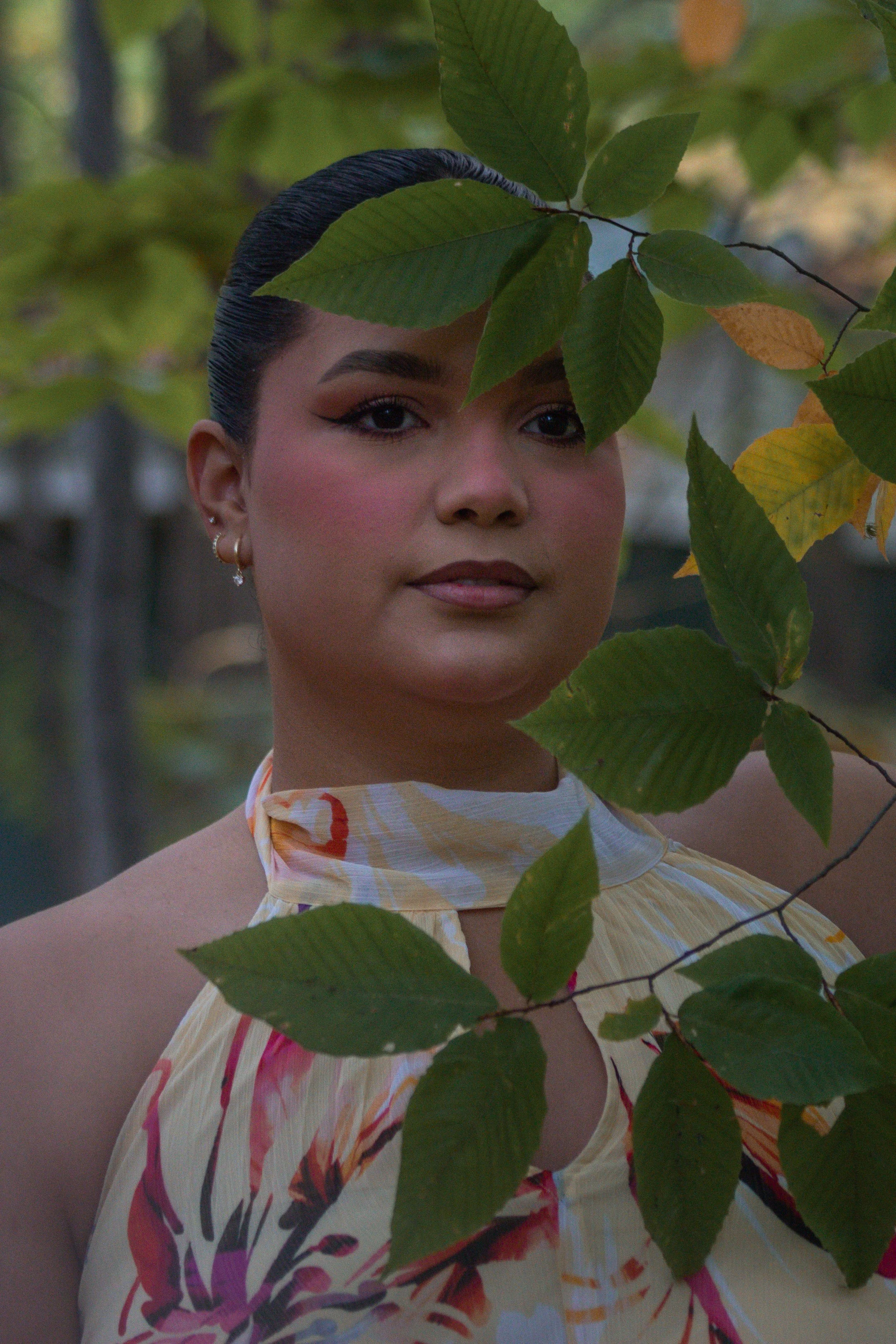 A woman with dark hair, makeup, and earrings is partially obscured by green leaves and yellowing leaves, with a blurred outdoor background.