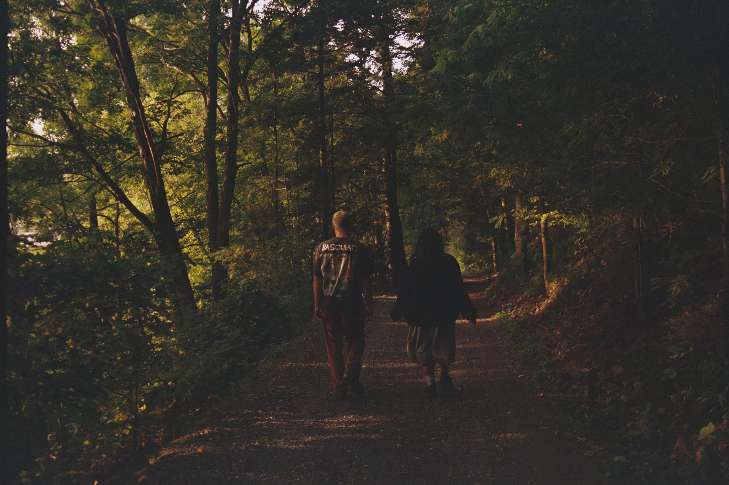 Three people walking on a forest trail during dusk, surrounded by tall trees and dense greenery.