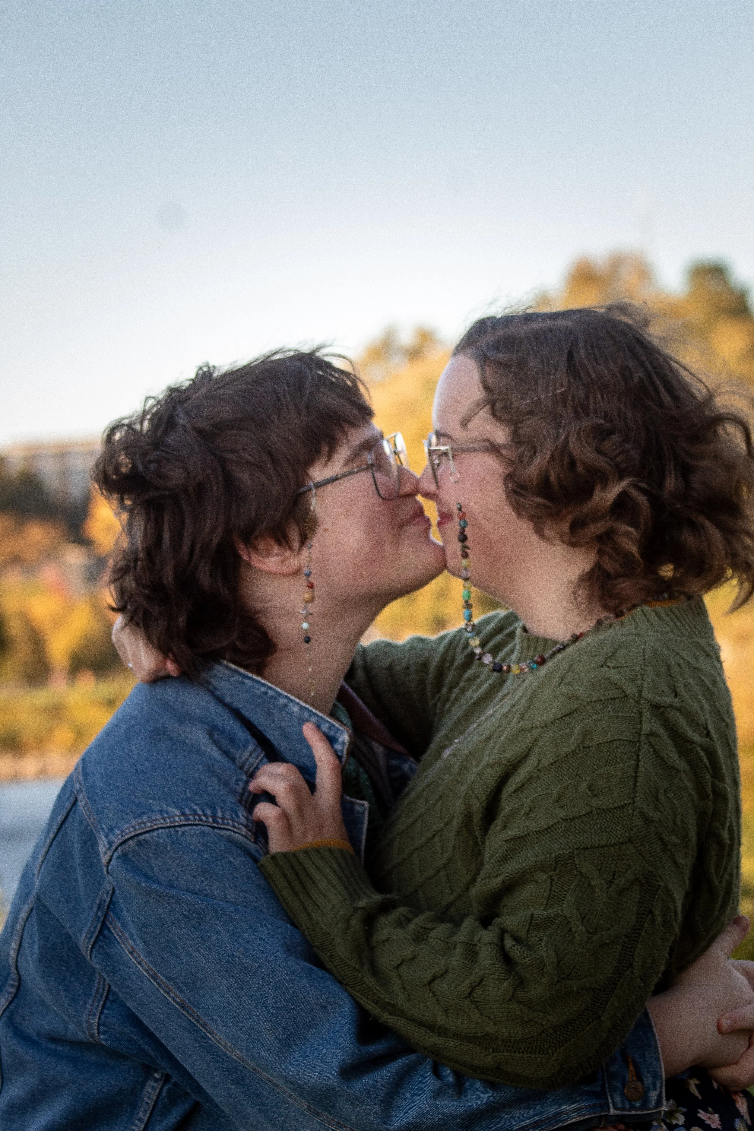 Two people with short, curly hair and glasses kissing outdoors during sunset, with trees in the background.