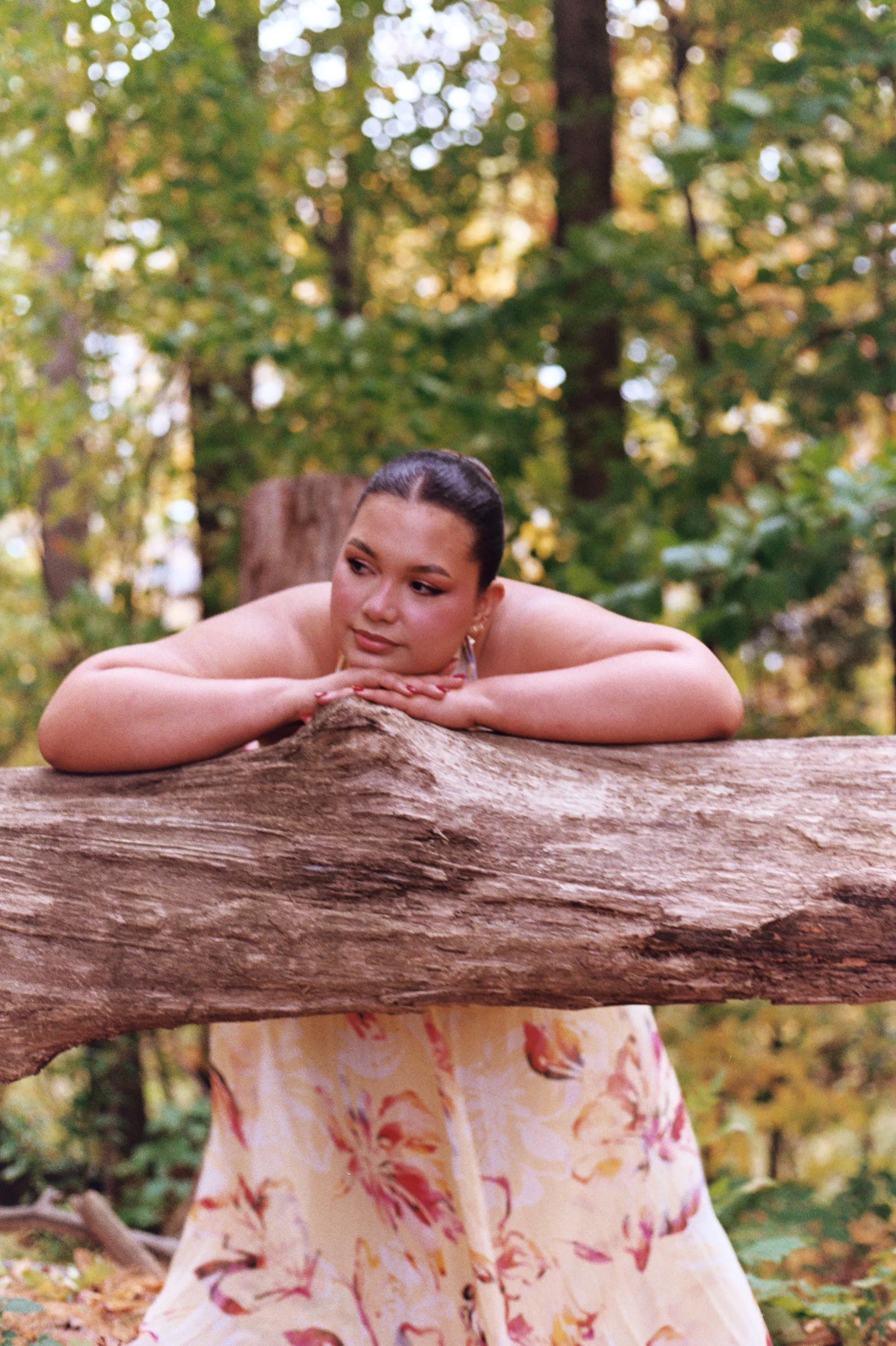 A woman with dark hair leaning on a large fallen tree trunk in a forest, with a woman in a colorful dress partially obscured behind the trunk. The background is filled with green leaves and trees.