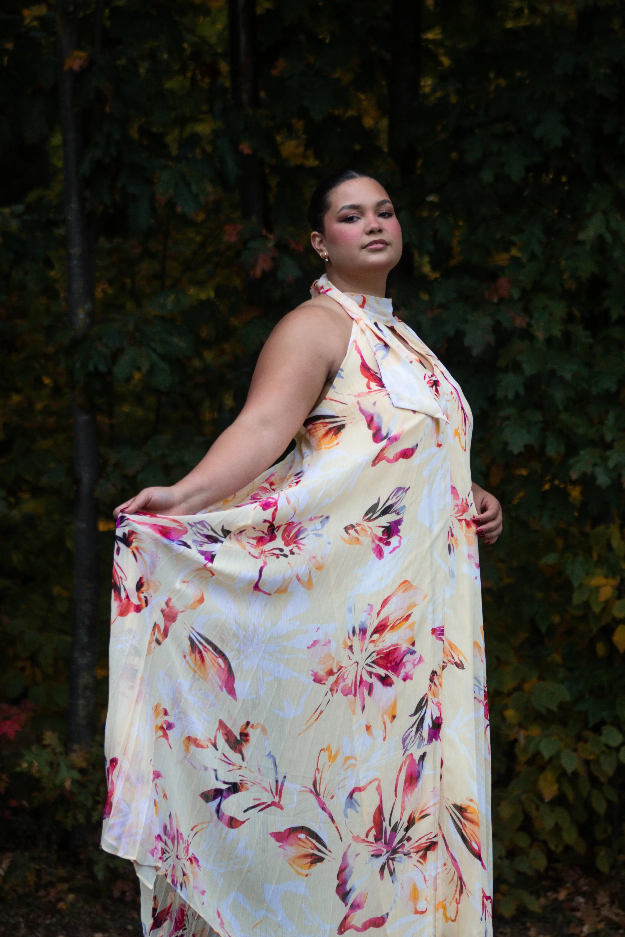 A woman with dark hair, wearing a sleeveless floral dress, stands outdoors in front of a background of green trees with some autumn leaves.