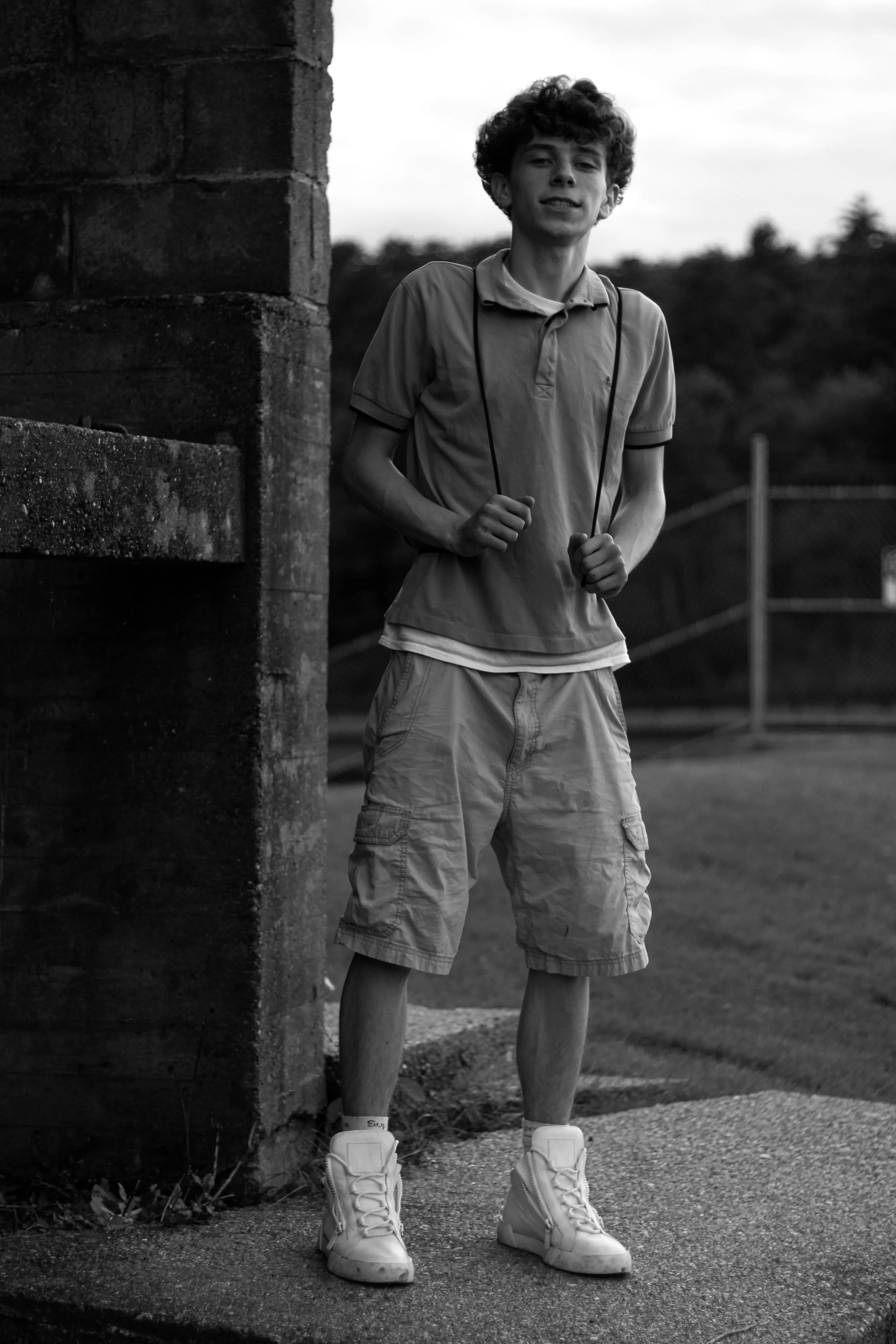 Black and white photo of a young man with curly hair standing outdoors behind a brick wall, smiling. He is dressed in a polo shirt, shorts, and high-top sneakers, with a lanyard around his neck. The background includes a fence and trees.