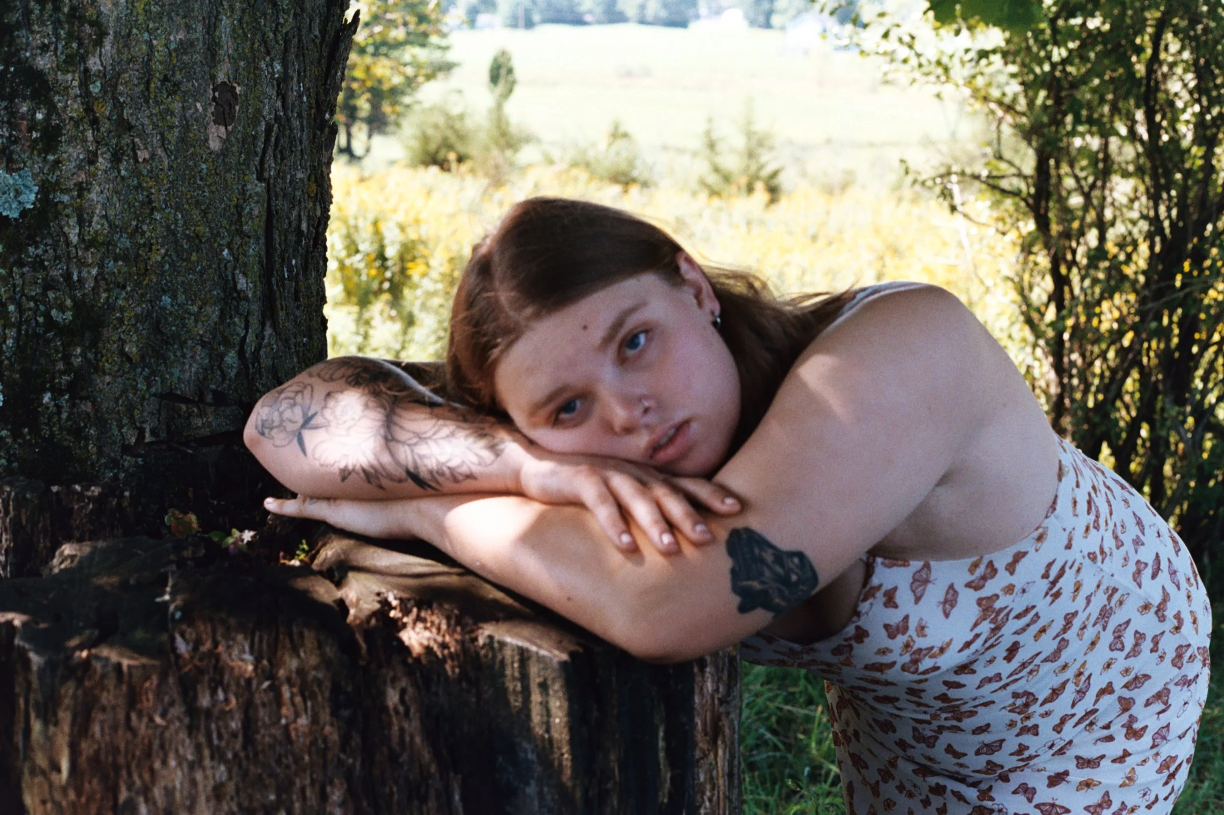 A woman with tattoos lying on a tree trunk in a forest, resting her head on her arms, with trees and sunlight in the background.