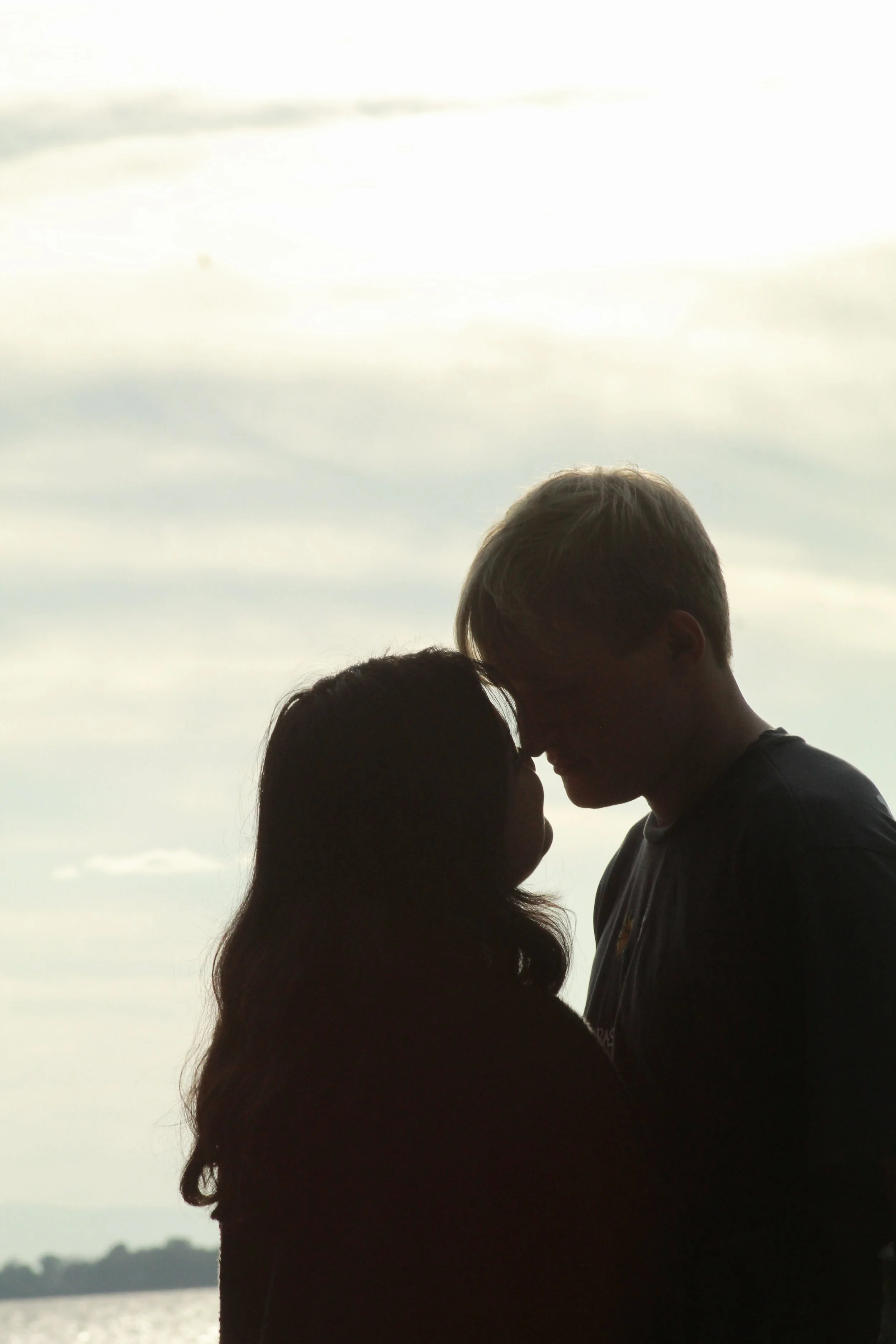 Silhouettes of a man and woman close together facing each other on a beach during sunset or sunrise, with water and distant land in the background.