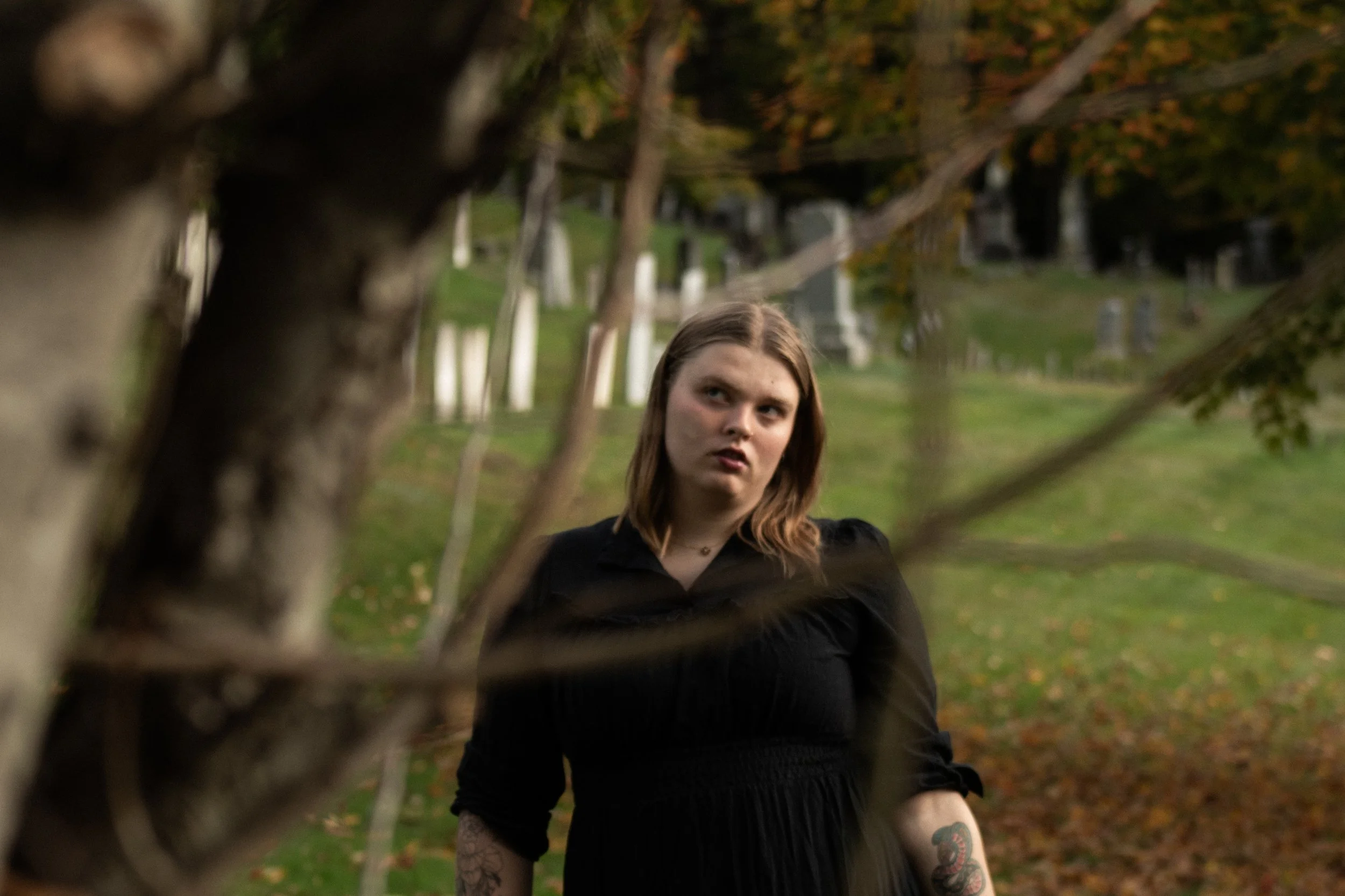 A woman with light brown hair and tattoos on her arm is standing outdoors in a park, looking through a tree trunk. The background features a grassy area with trees and tombstones, suggesting a cemetery setting.