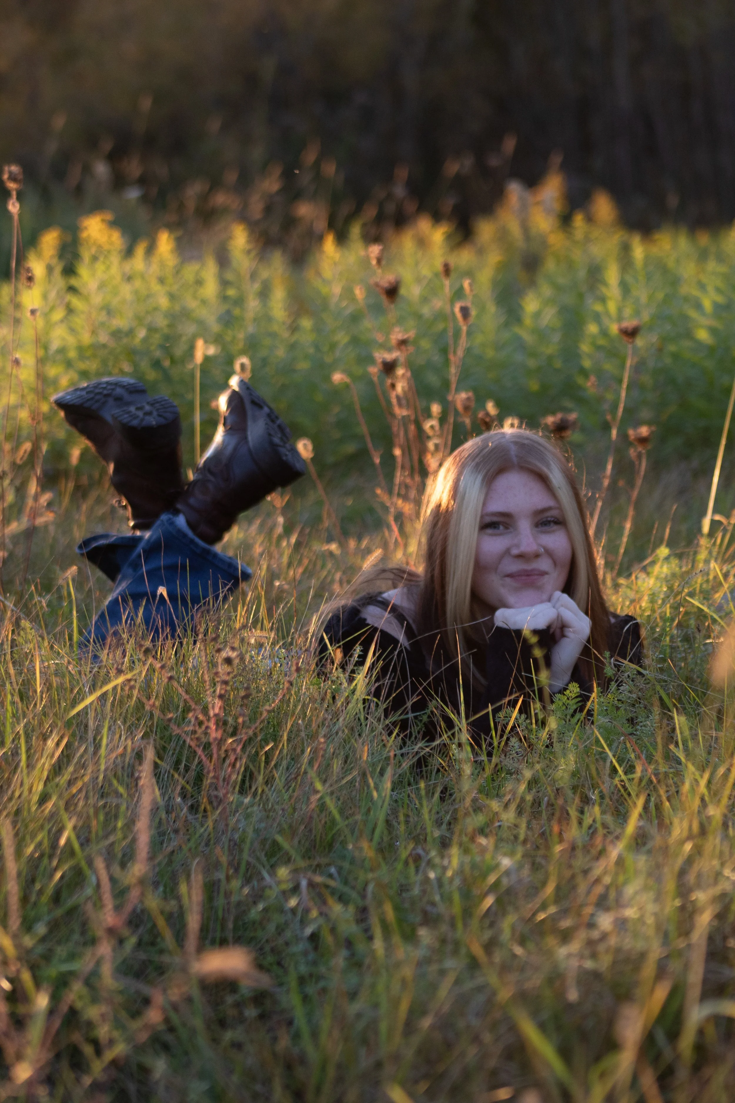 A young woman with blonde hair lying on green grass in a field, with one person lying upside down with their feet in the air in the background, during sunset.