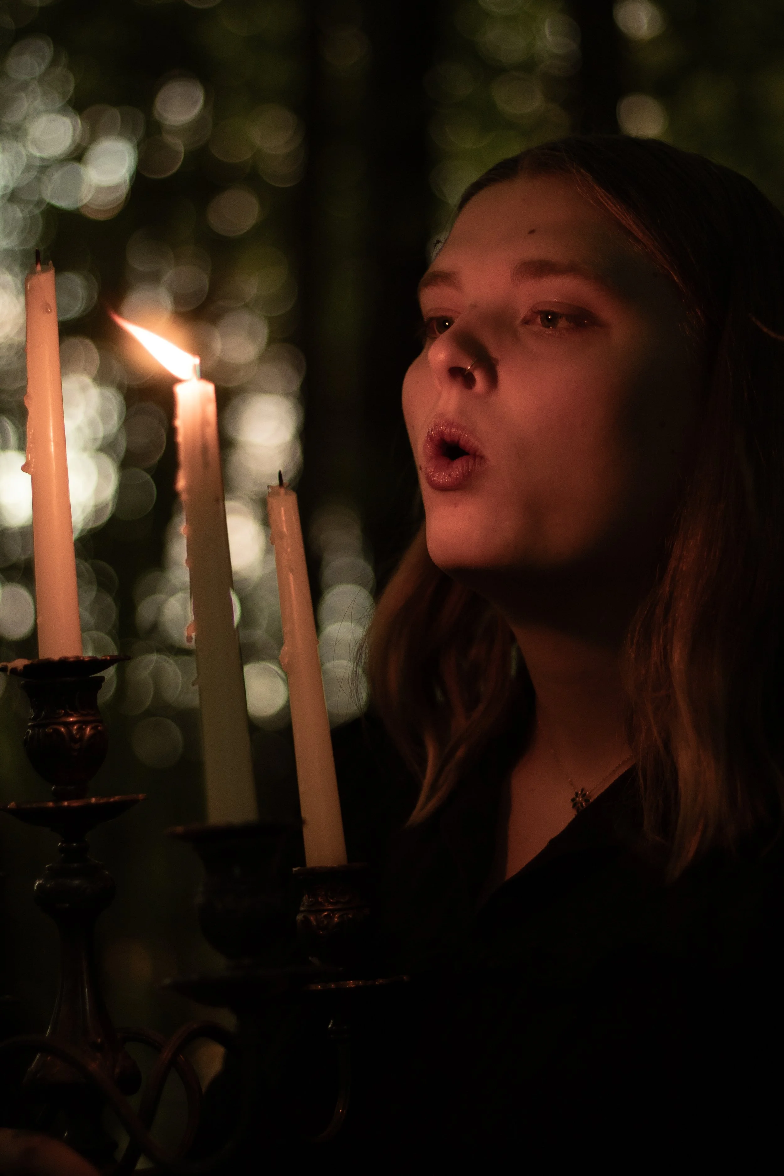 A young woman with long brown hair and a nose piercing, illuminated by candlelight, in front of a dark background with blurred light bokeh.