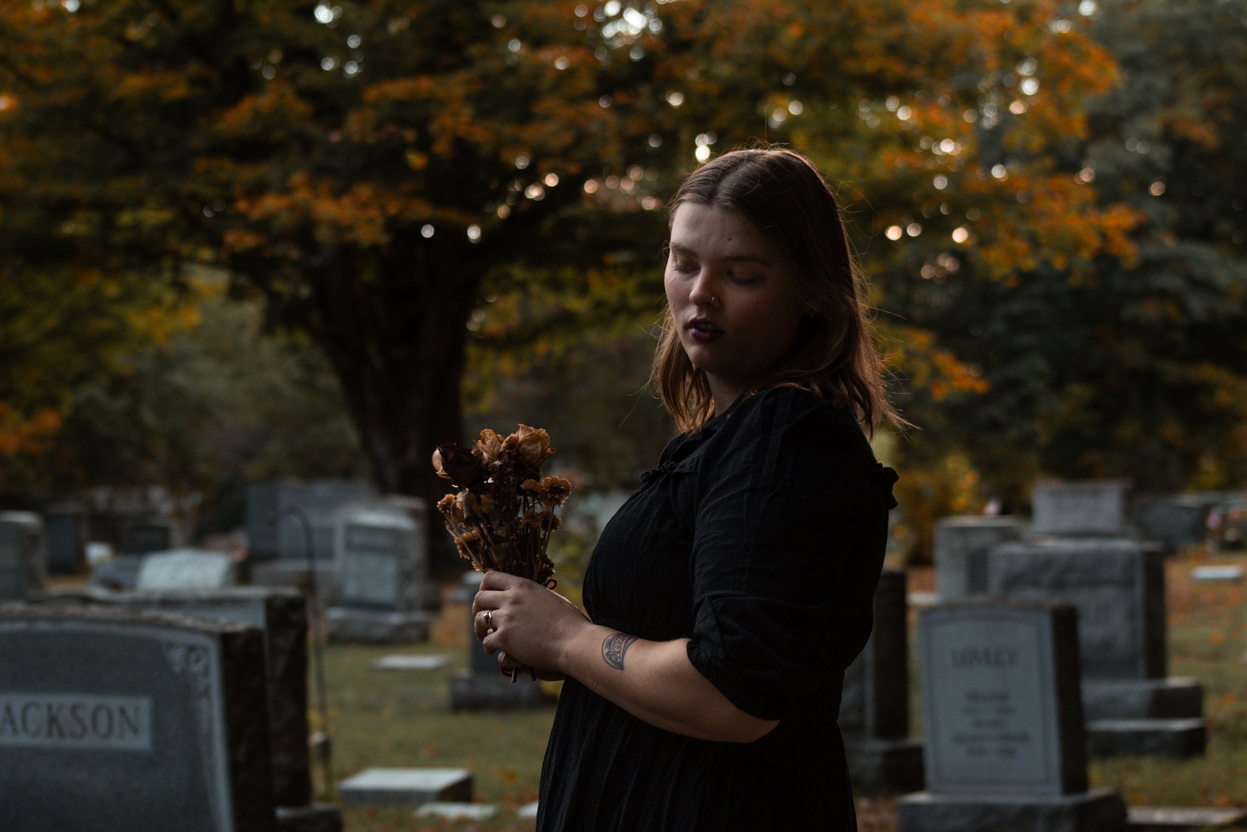 A woman with long brown hair, wearing a black jacket, holding a bouquet of dried flowers, standing in a cemetery with headstones and a large tree with orange and yellow leaves in the background, during autumn.