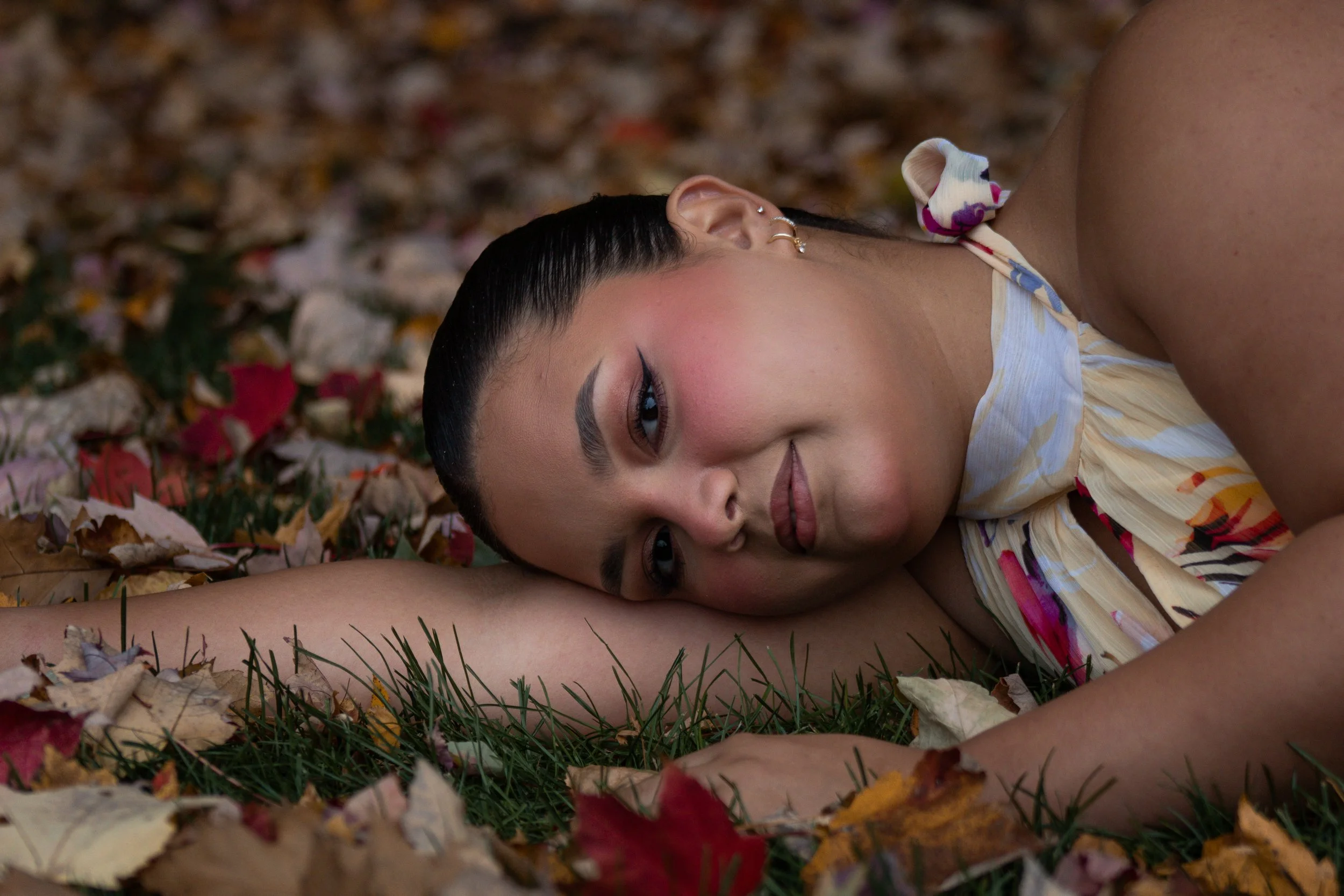A woman lying on the ground among autumn leaves smiling, with dry grass and colorful leaves scattered around.