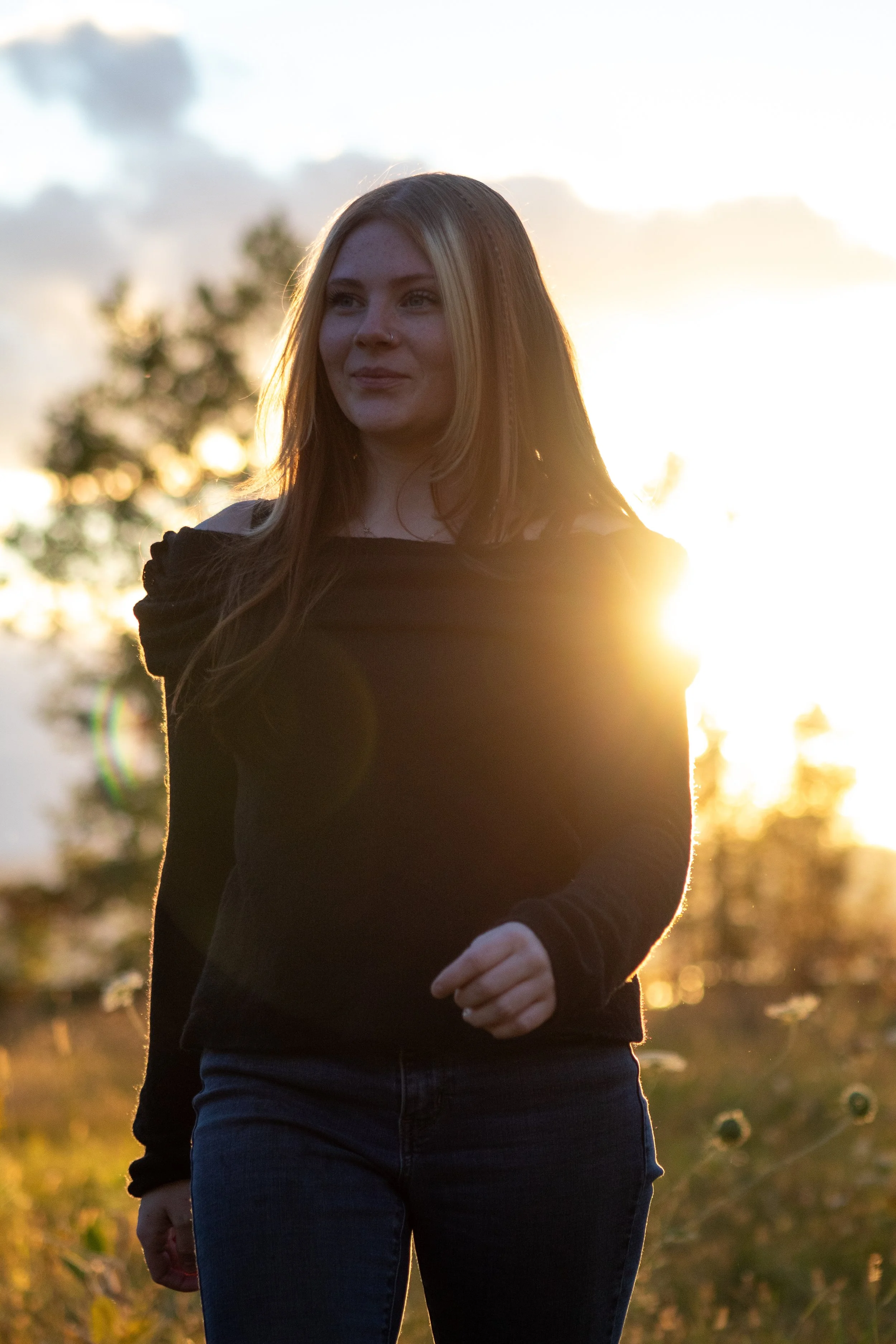 A woman with long light brown hair wearing a black long sleeve top and jeans, standing outdoors during sunset with trees and sky in the background.