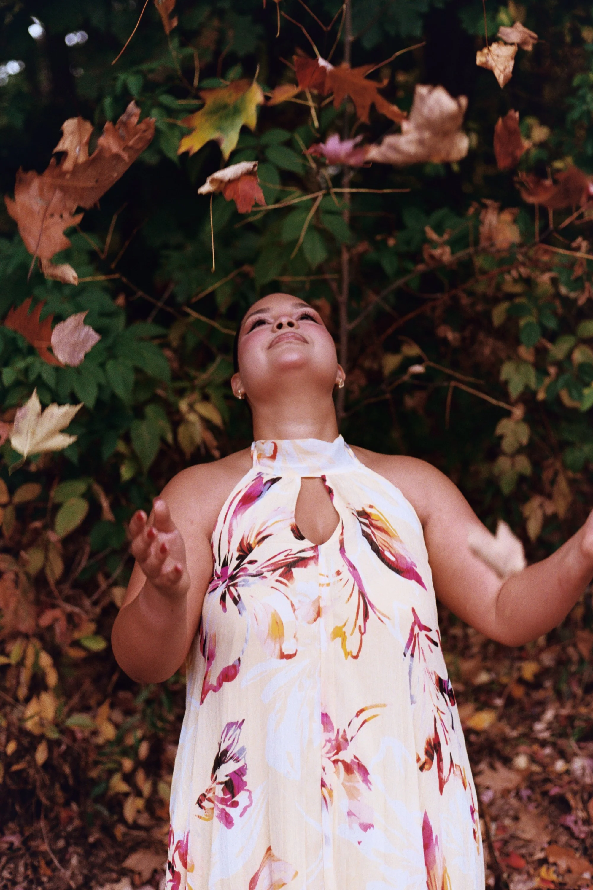 A woman outdoors surrounded by autumn leaves, looking up with a joyful expression, wearing a floral dress.