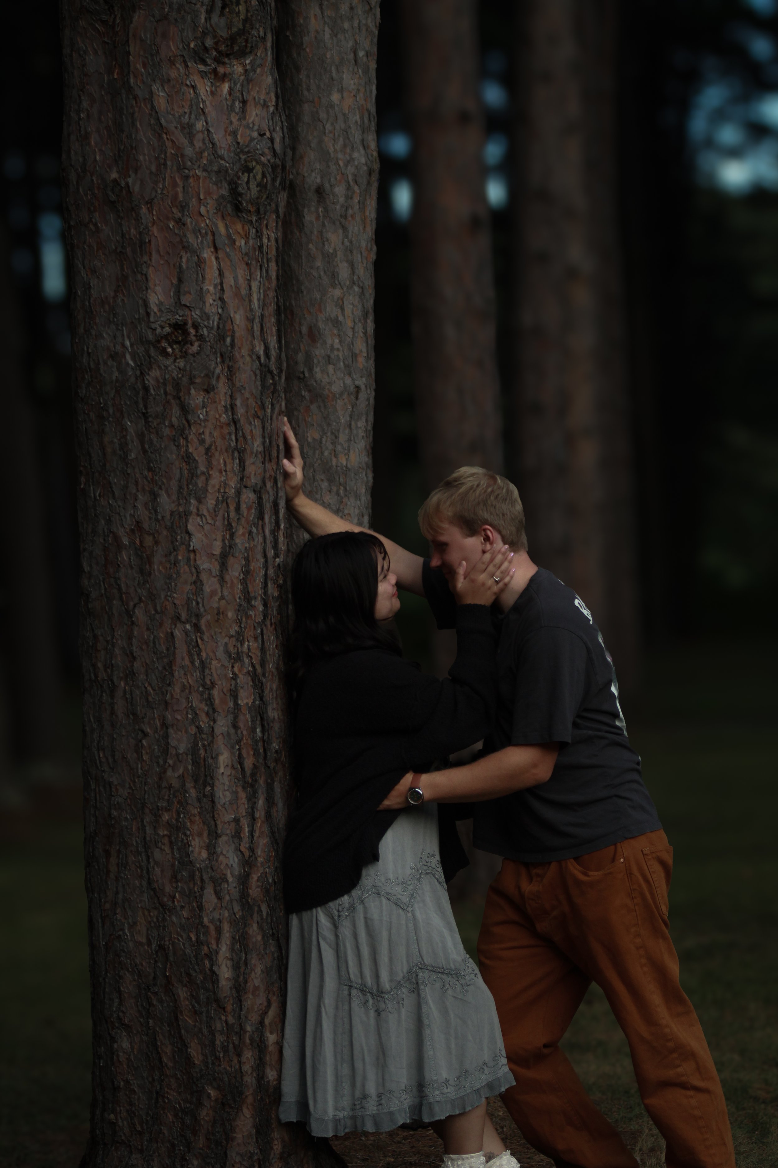 A couple stands close to a large tree in a forest, looking into each other's eyes, with their hands gently touching each other's faces.